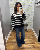 A woman takes a mirror selfie wearing a black and white horizontally striped knit sweater, blue flare jeans, and tan boots in a fitting room with wood floors and abstract wallpaper.