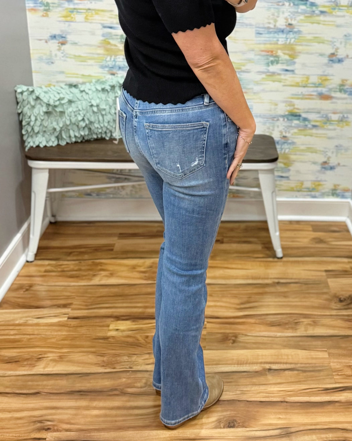 A person wearing mica bootcut jeans, a black top, and tan boots, standing in a room with wood floors and a bench in the background.