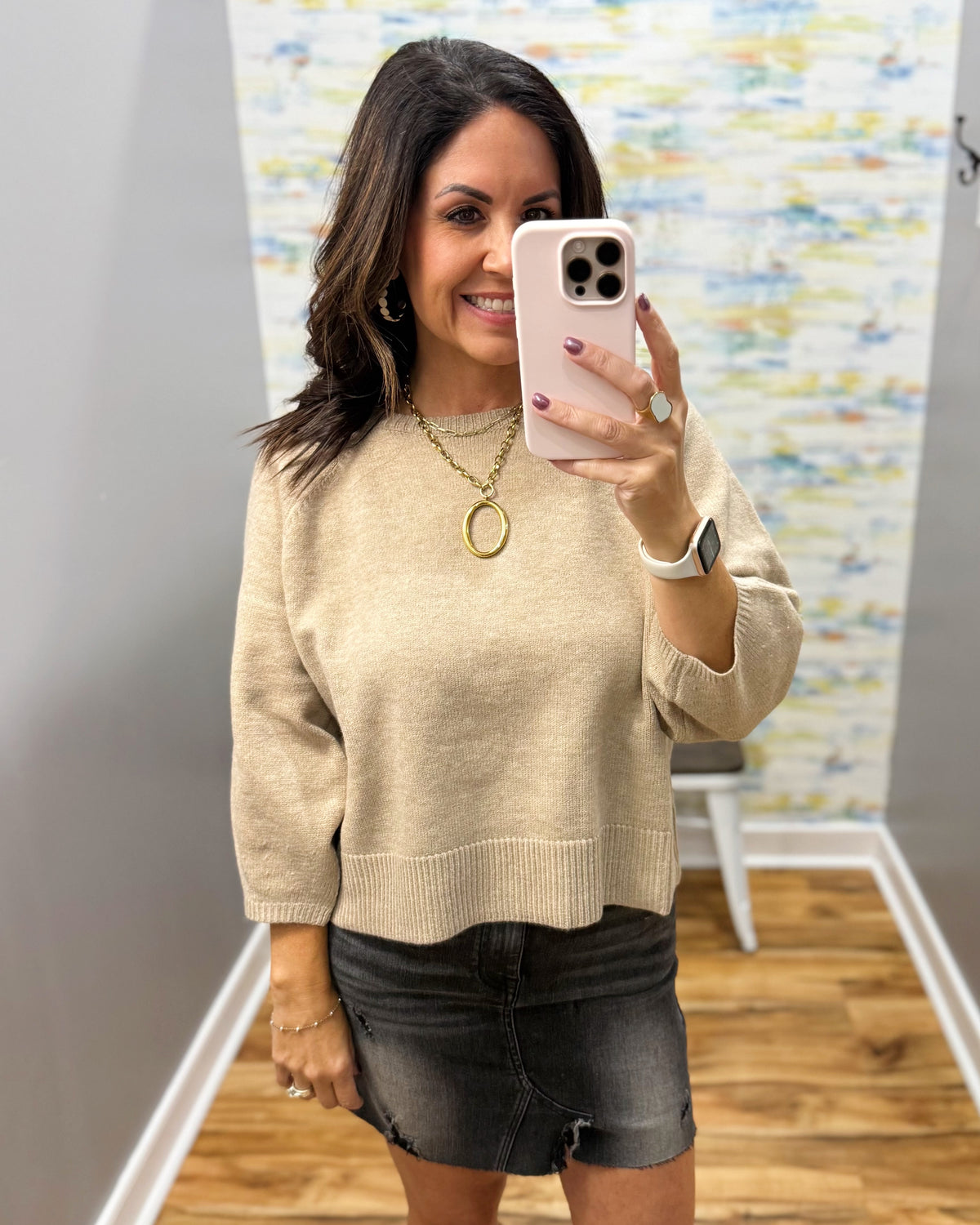 A woman takes a mirror selfie wearing a beige knit sweater and a black denim mini skirt in what appears to be a home or boutique fitting space.