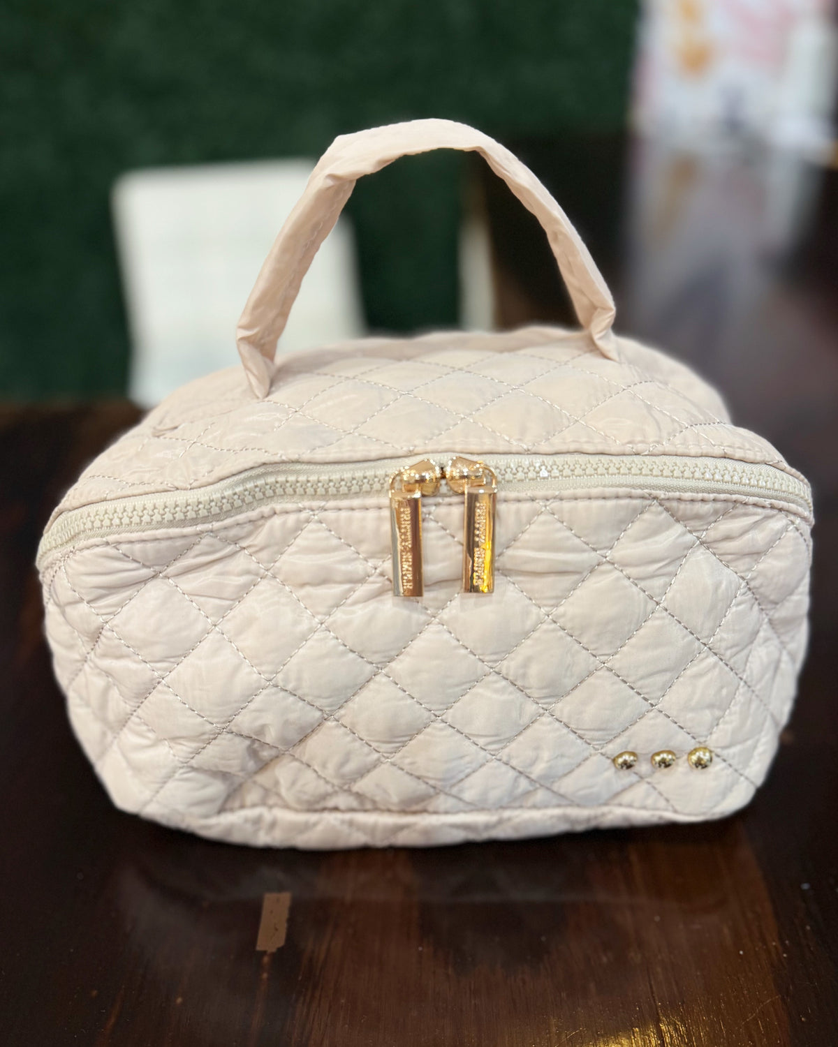 A quilted taupe-colored cosmetic bag with a top handle, gold zipper, and gold hardware, resting on a dark wooden table.
