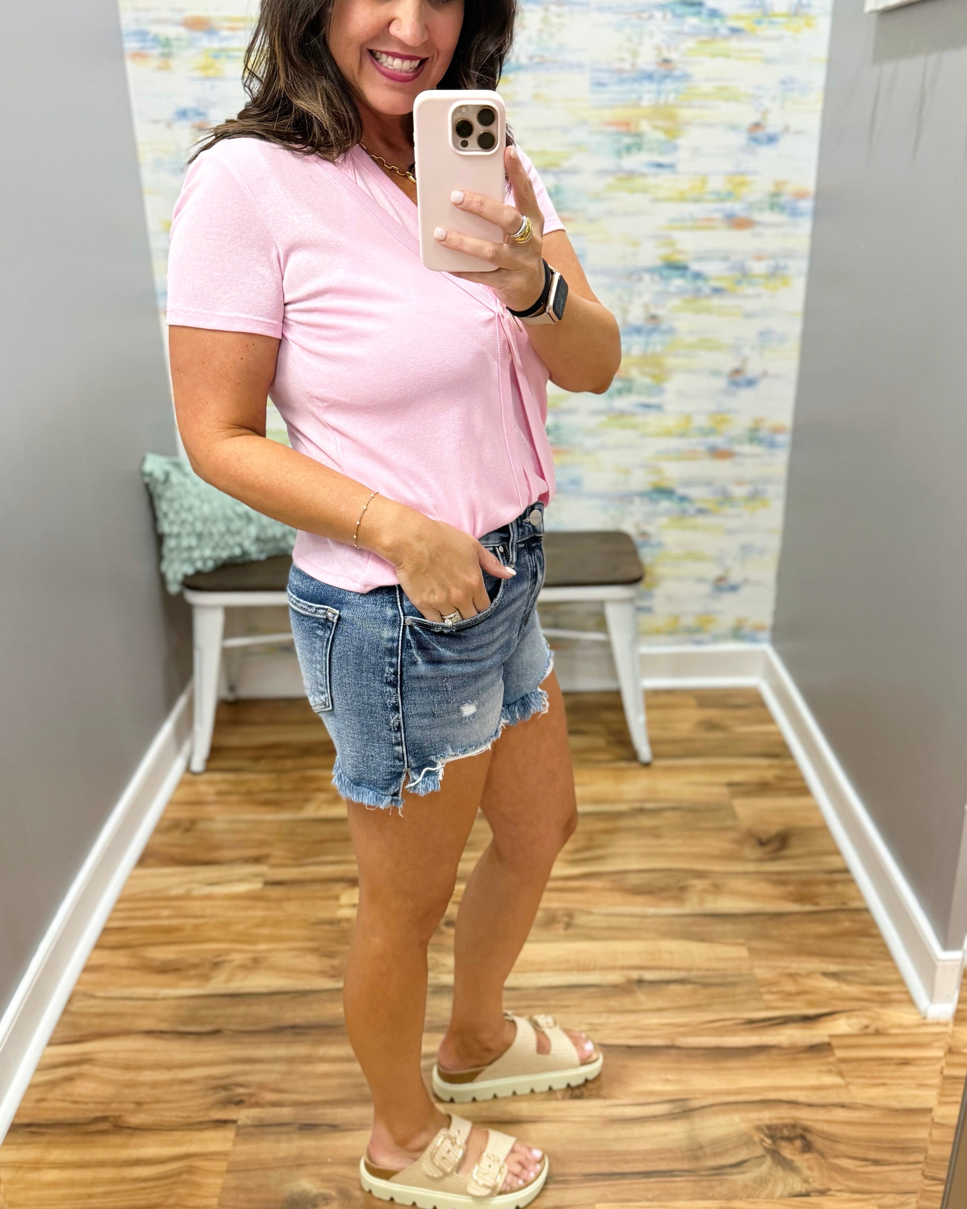 Woman taking a mirror selfie wearing a pink t-shirt and denim shorts in a room with a colorful wall.