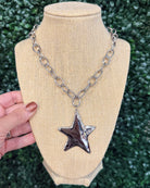 A silver-toned chunky chain necklace with a textured, hammered star-shaped pendant, displayed on a beige burlap-covered jewelry bust. A hand with glittery nail polish is holding the left side of the bust, and a green hedge wall serves as the background.