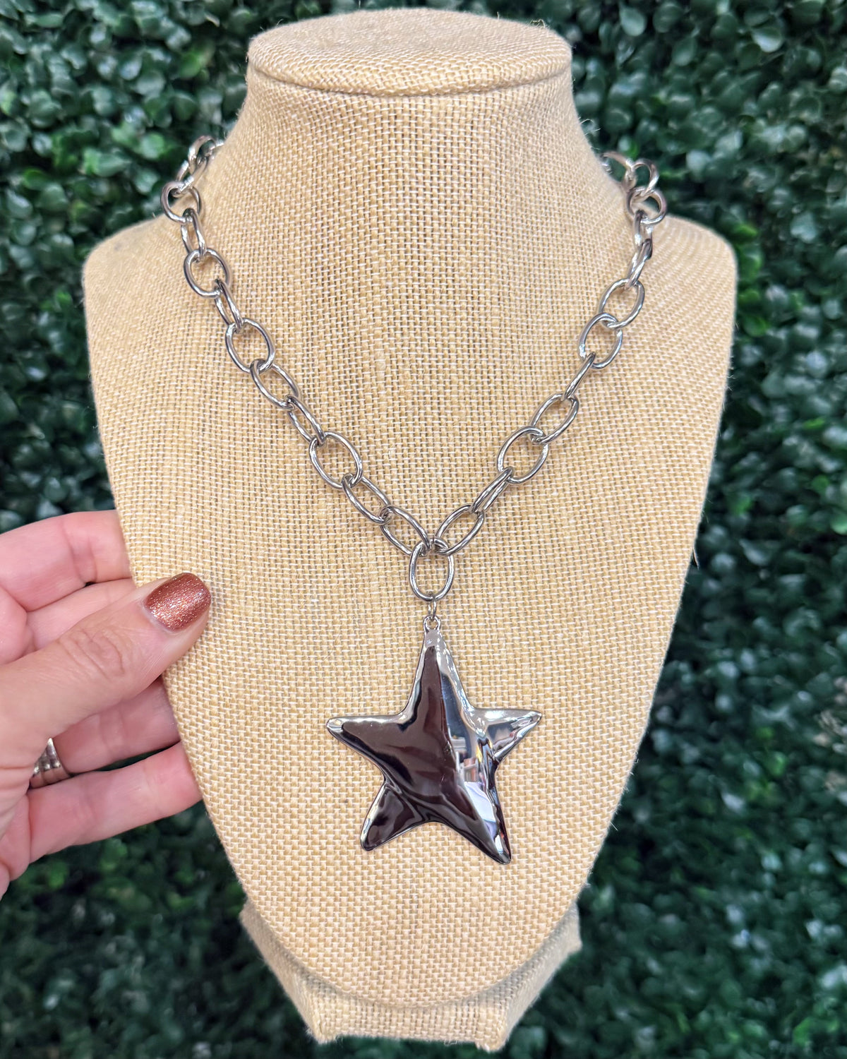 A silver-toned chunky chain necklace with a textured, hammered star-shaped pendant, displayed on a beige burlap-covered jewelry bust. A hand with glittery nail polish is holding the left side of the bust, and a green hedge wall serves as the background.