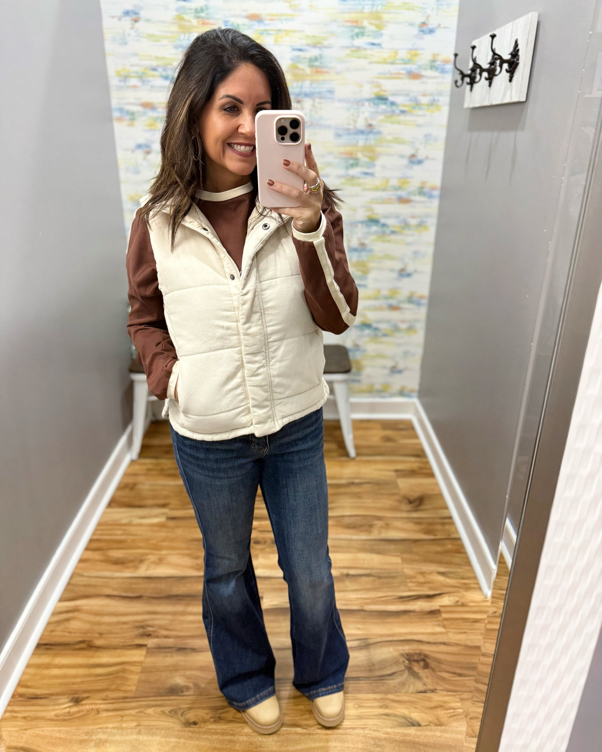 A woman takes a mirror selfie showing off a cream puffer vest with brown sleeves, worn over a brown top and blue jeans.
