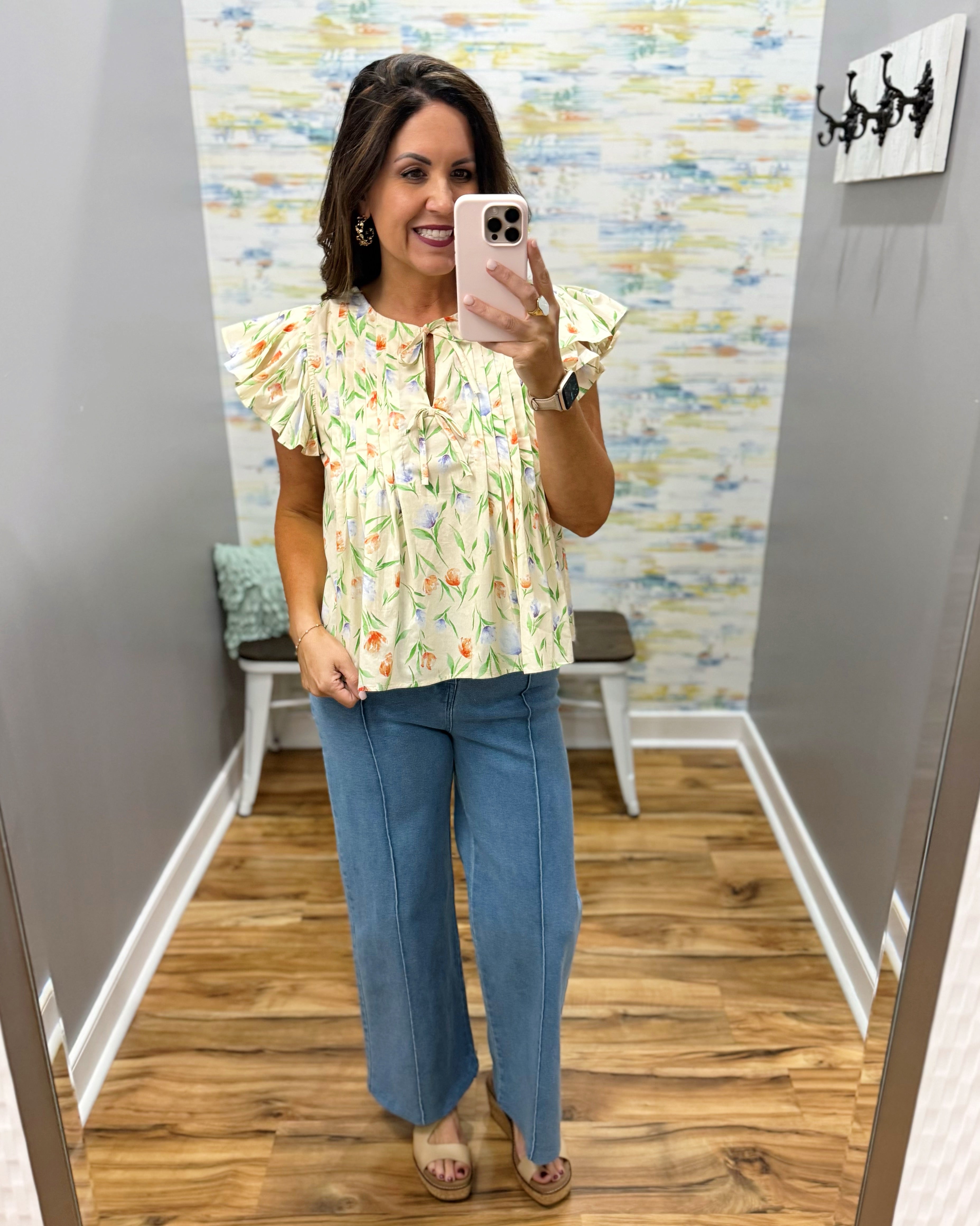 Woman taking a mirror selfie wearing a floral blouse and blue jeans in a room with a colorful wall.