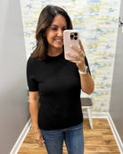 A woman taking a mirror selfie wearing a black short-sleeve crewneck top and blue jeans, holding a light pink smartphone.