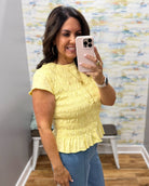 A woman taking a mirror selfie wearing a light yellow short-sleeve ruffled blouse and blue jeans in a fitting room with a patterned wall.