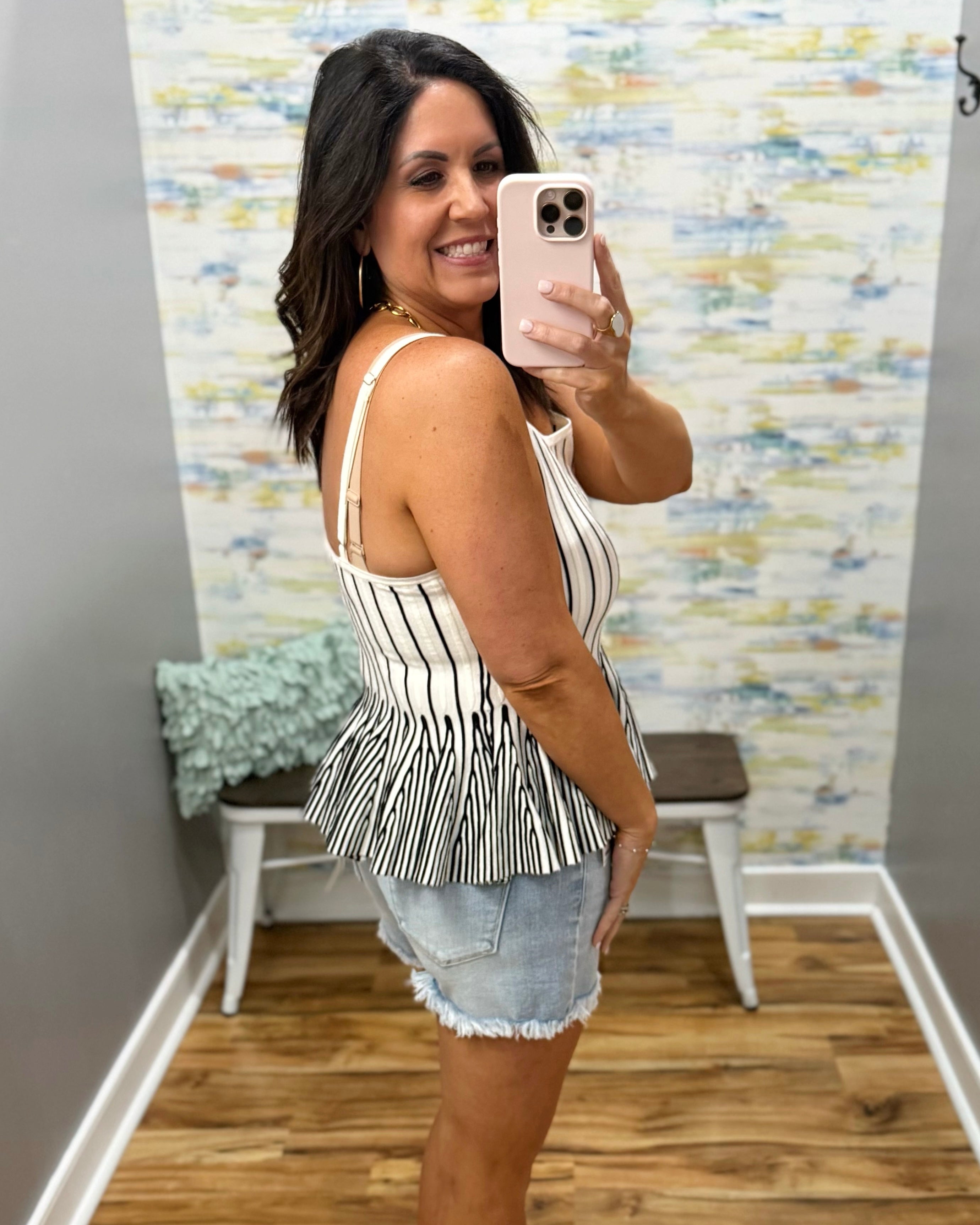 Woman taking a mirror selfie wearing a striped peplum top and denim shorts in a room with patterned wallpaper.