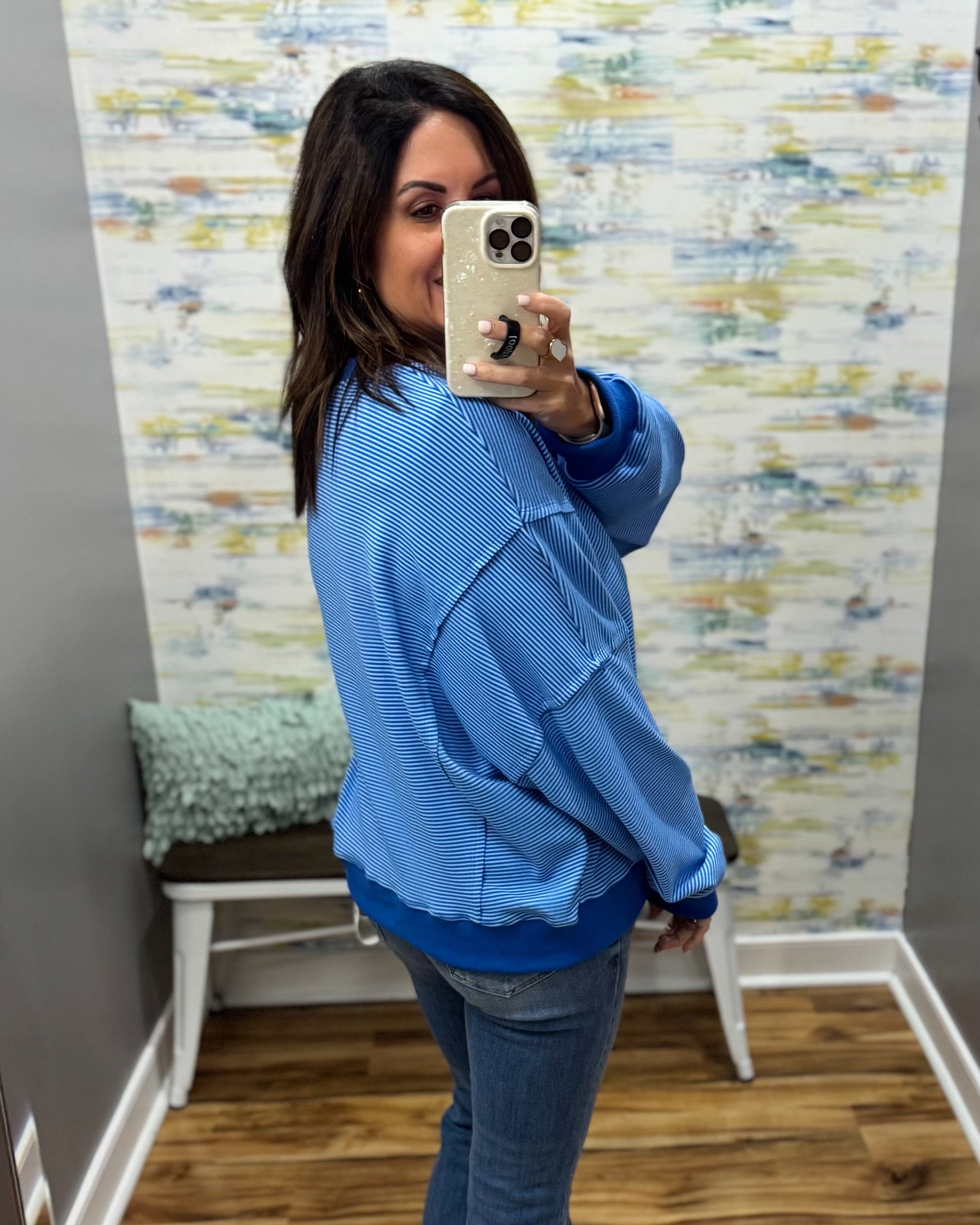 A woman takes a mirror selfie wearing a light blue textured waffle-knit sweatshirt and blue jeans in a room with patterned wallpaper and a wooden floor.