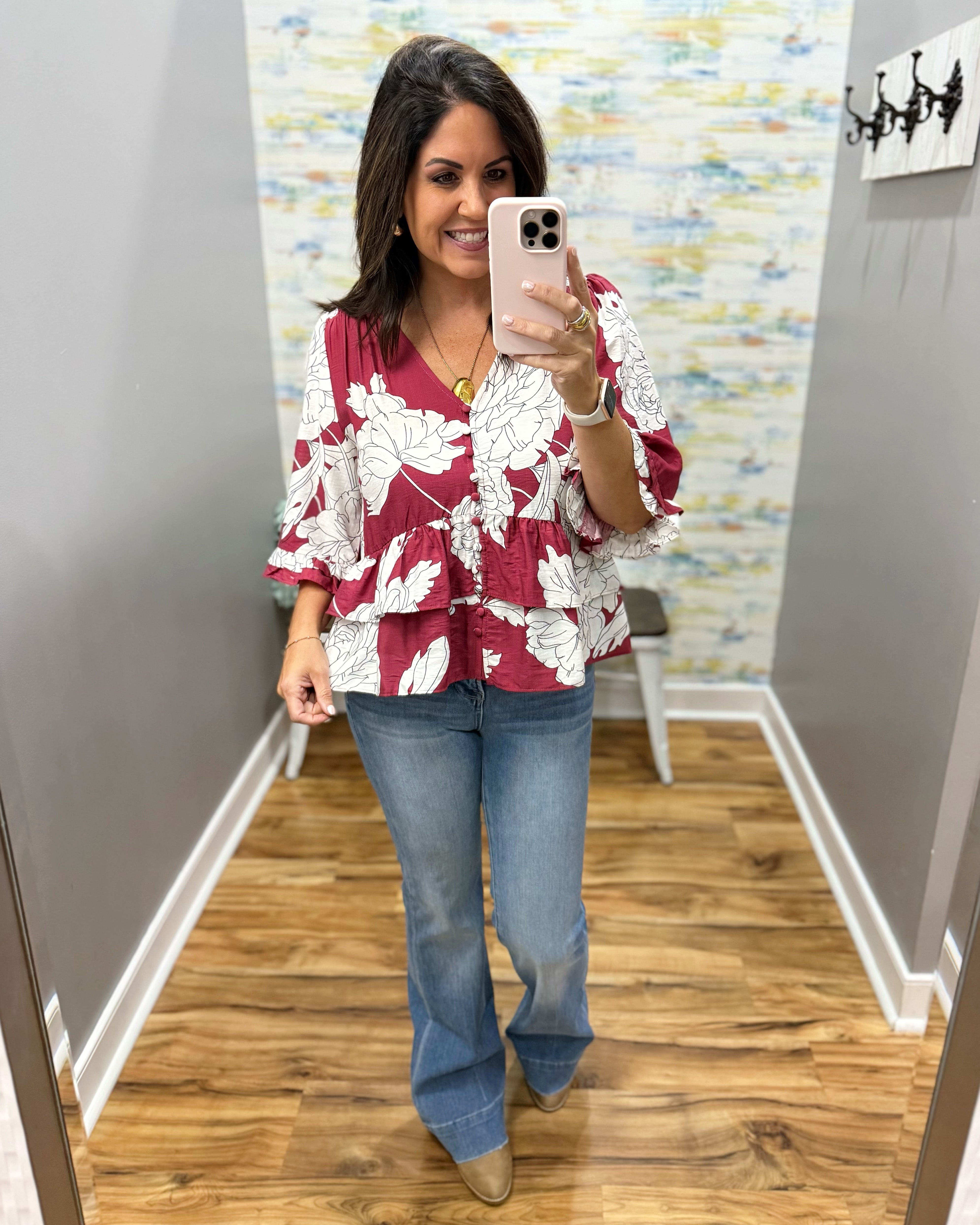 A woman takes a mirror selfie in a fitting room wearing a burgundy and white floral print peplum blouse, blue jeans, and tan boots.