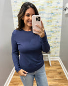 A woman taking a mirror selfie in a fitting room wearing a navy blue long-sleeve crewneck shirt and light wash jeans.