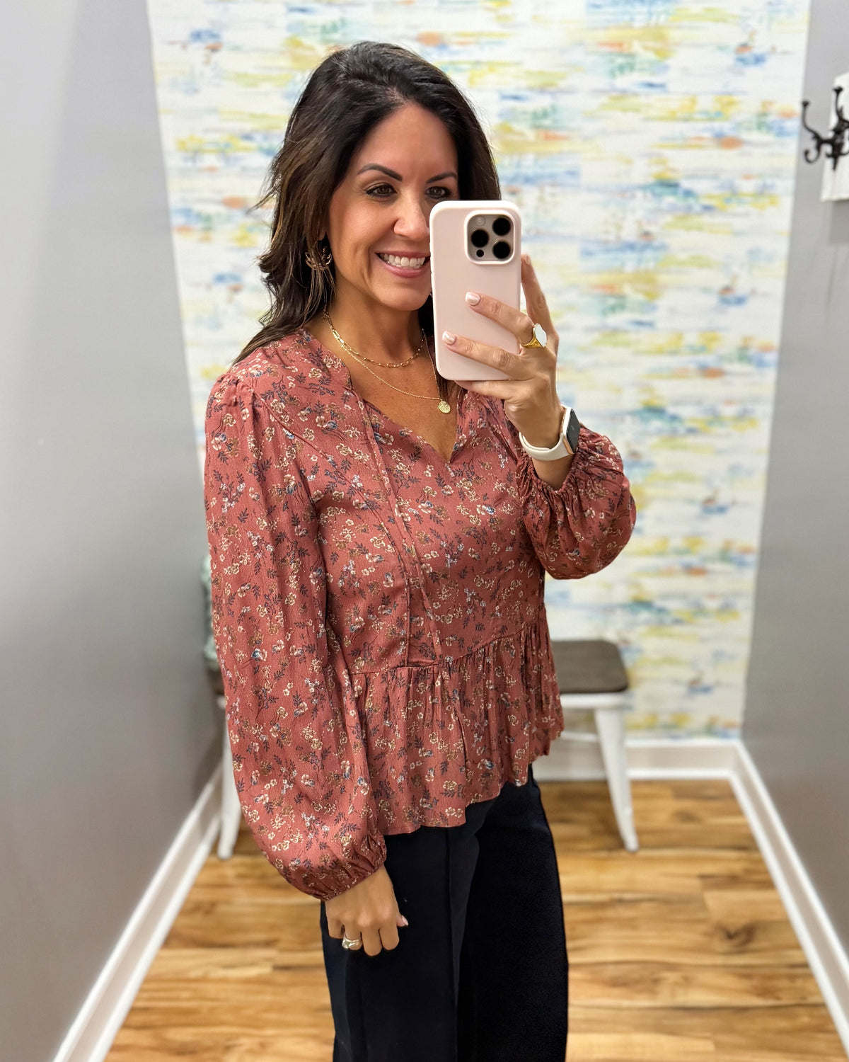 A woman takes a mirror selfie wearing a dusty rose long-sleeve blouse with a small white floral pattern and dark pants, standing in a fitting room with a wood floor and abstract wall art.
