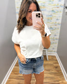 Woman taking a mirror selfie wearing a white t-shirt and denim shorts in a room with a patterned wall.