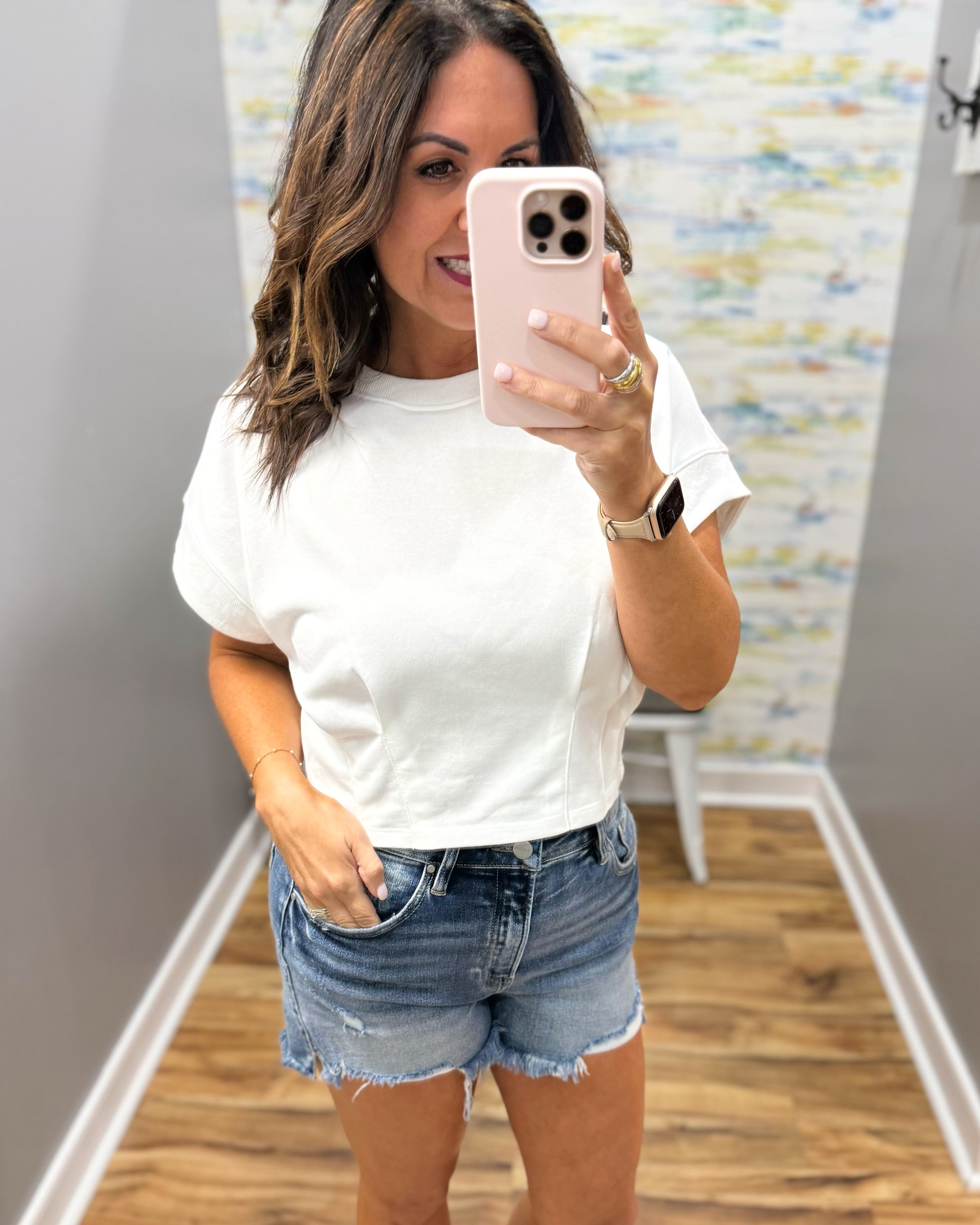 Woman taking a mirror selfie wearing a white t-shirt and denim shorts in a room with a patterned wall.