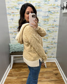 A woman takes a mirror selfie in a fitting room wearing a textured, heathered cream and tan hooded sweater with ribbed trim, paired with blue jeans.