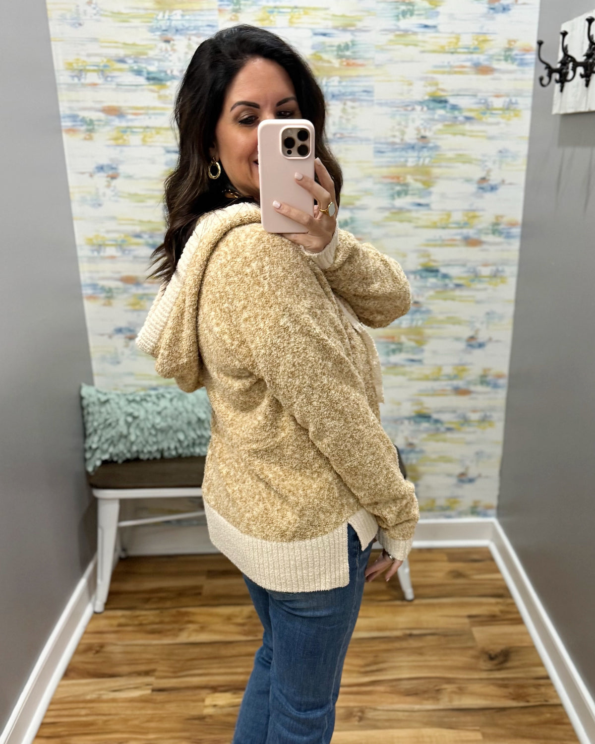 A woman takes a mirror selfie in a fitting room wearing a textured, heathered cream and tan hooded sweater with ribbed trim, paired with blue jeans.