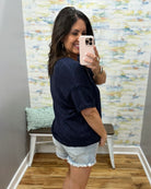 Woman taking a mirror selfie wearing a navy blue shirt and denim shorts in a room with a textured wall.