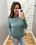 A woman taking a mirror selfie in a room, wearing a heathered sage green crewneck sweater and light blue jeans.