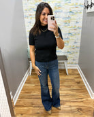 A woman takes a mirror selfie wearing a black short-sleeve mock neck top with scalloped edges and blue flare jeans, standing in a fitting room with wood floors and a patterned wallpaper background.