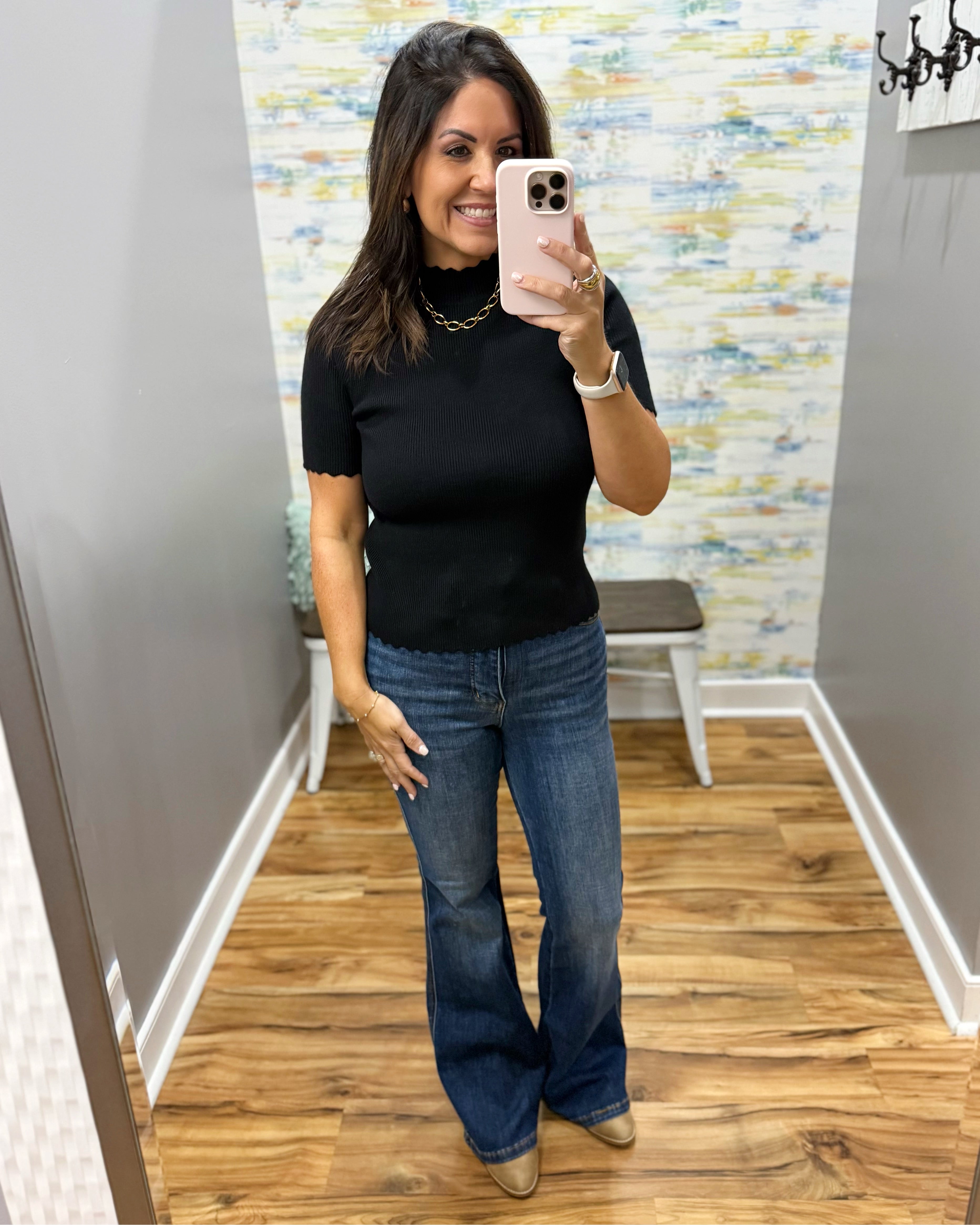 A woman takes a mirror selfie wearing a black short-sleeve mock neck top with scalloped edges and blue flare jeans, standing in a fitting room with wood floors and a patterned wallpaper background.