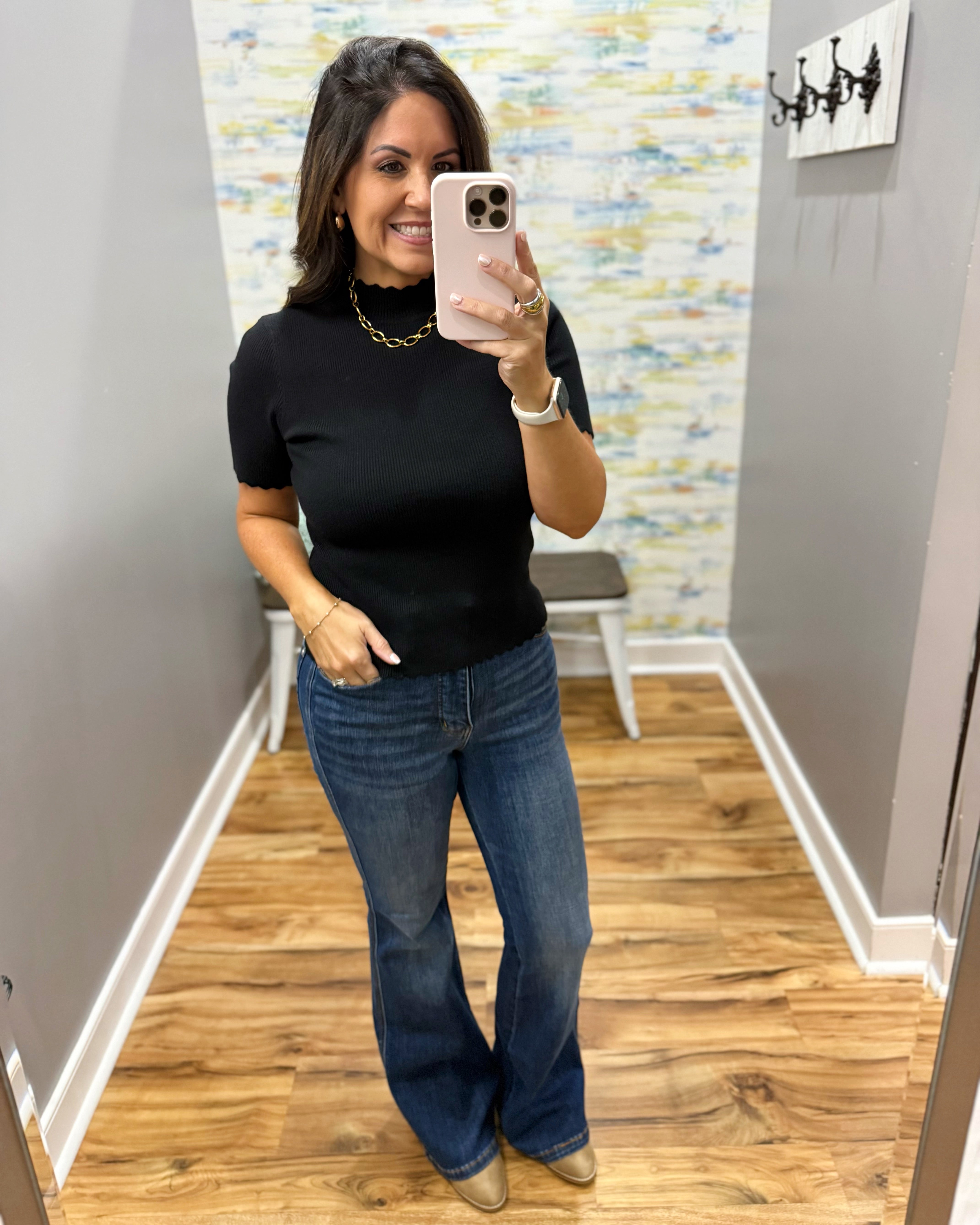A woman takes a mirror selfie in a fitting room wearing a black short-sleeve mock neck top and blue flare jeans.
