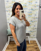 A woman takes a mirror selfie wearing a light gray knit short-sleeve top and dark blue jeans in a home setting with a patterned wallpaper background.