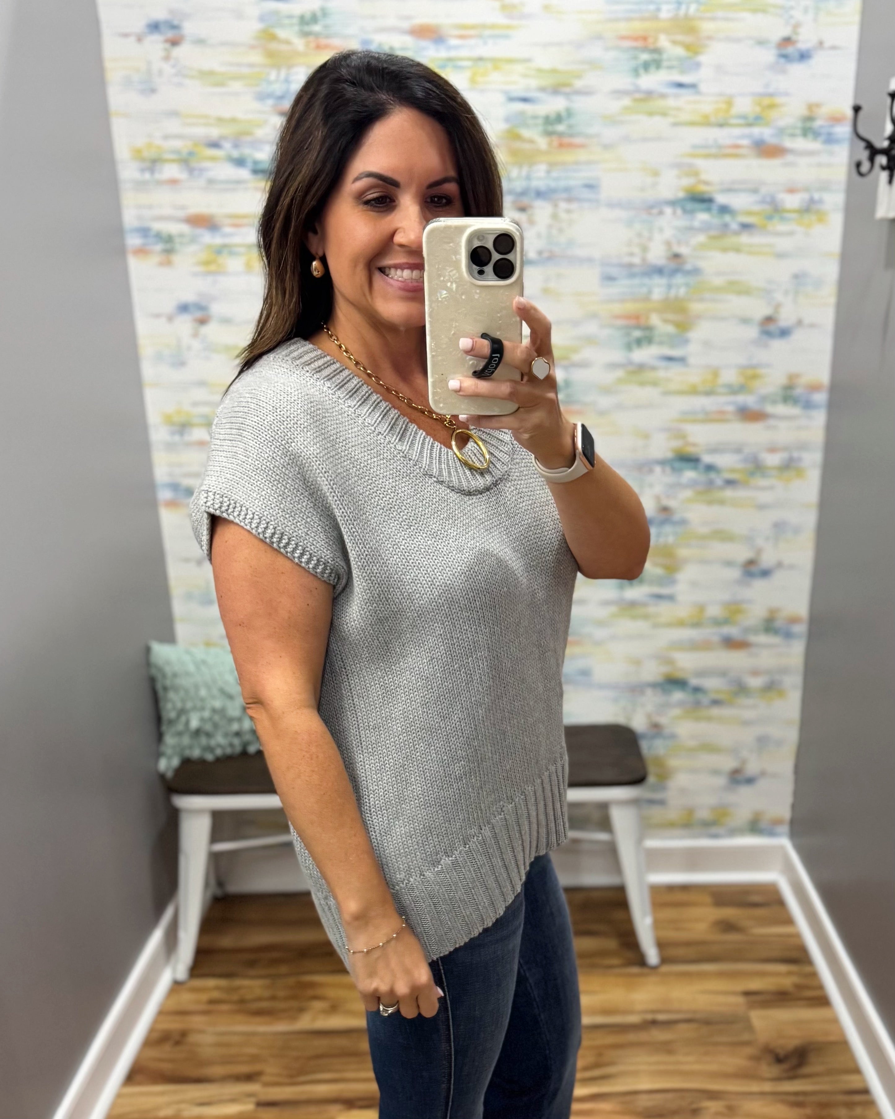 A woman takes a mirror selfie wearing a light gray knit short-sleeve top and dark blue jeans in a home setting with a patterned wallpaper background.