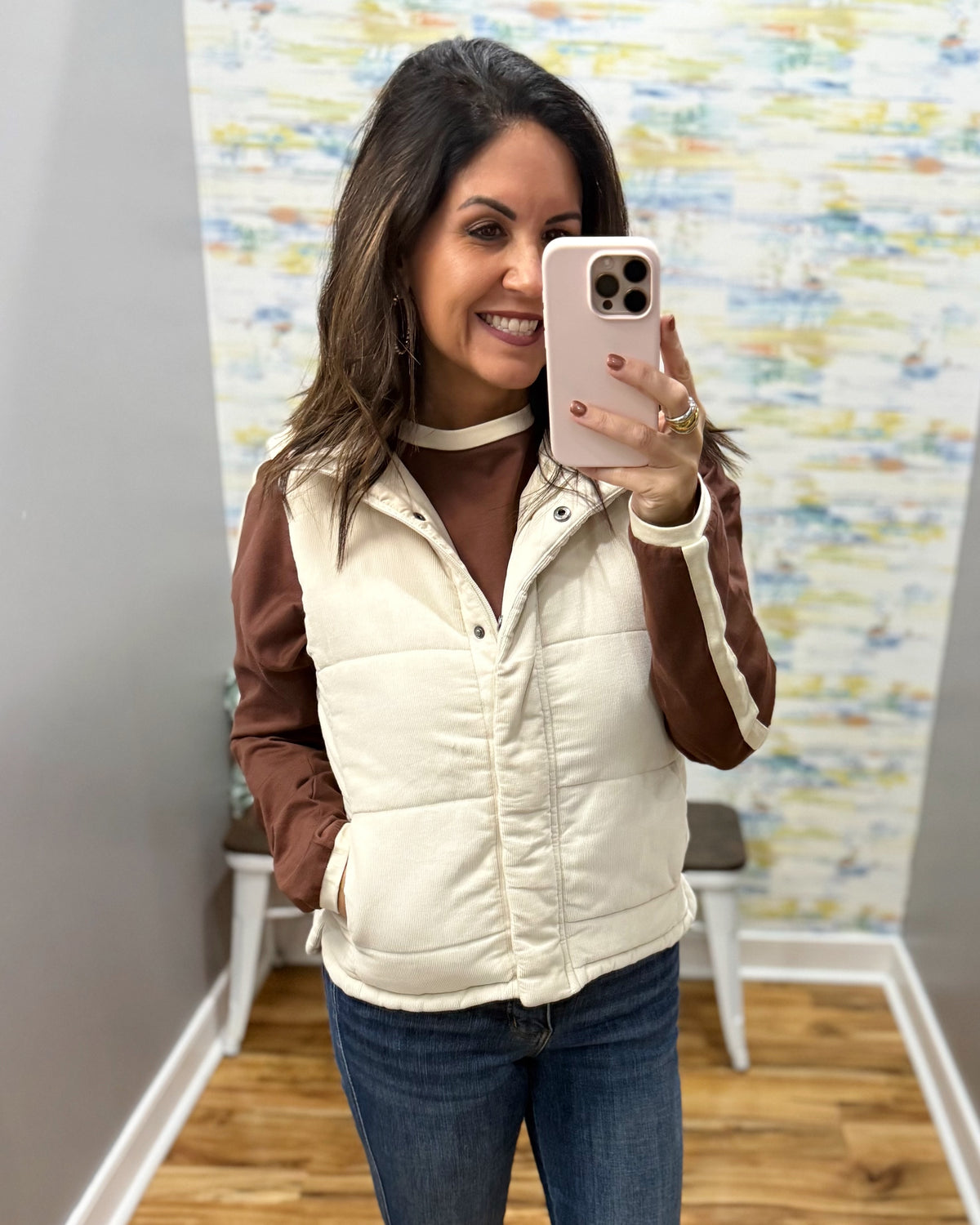 A woman takes a mirror selfie wearing a cream quilted puffer vest with brown sleeves over a brown top and blue jeans.