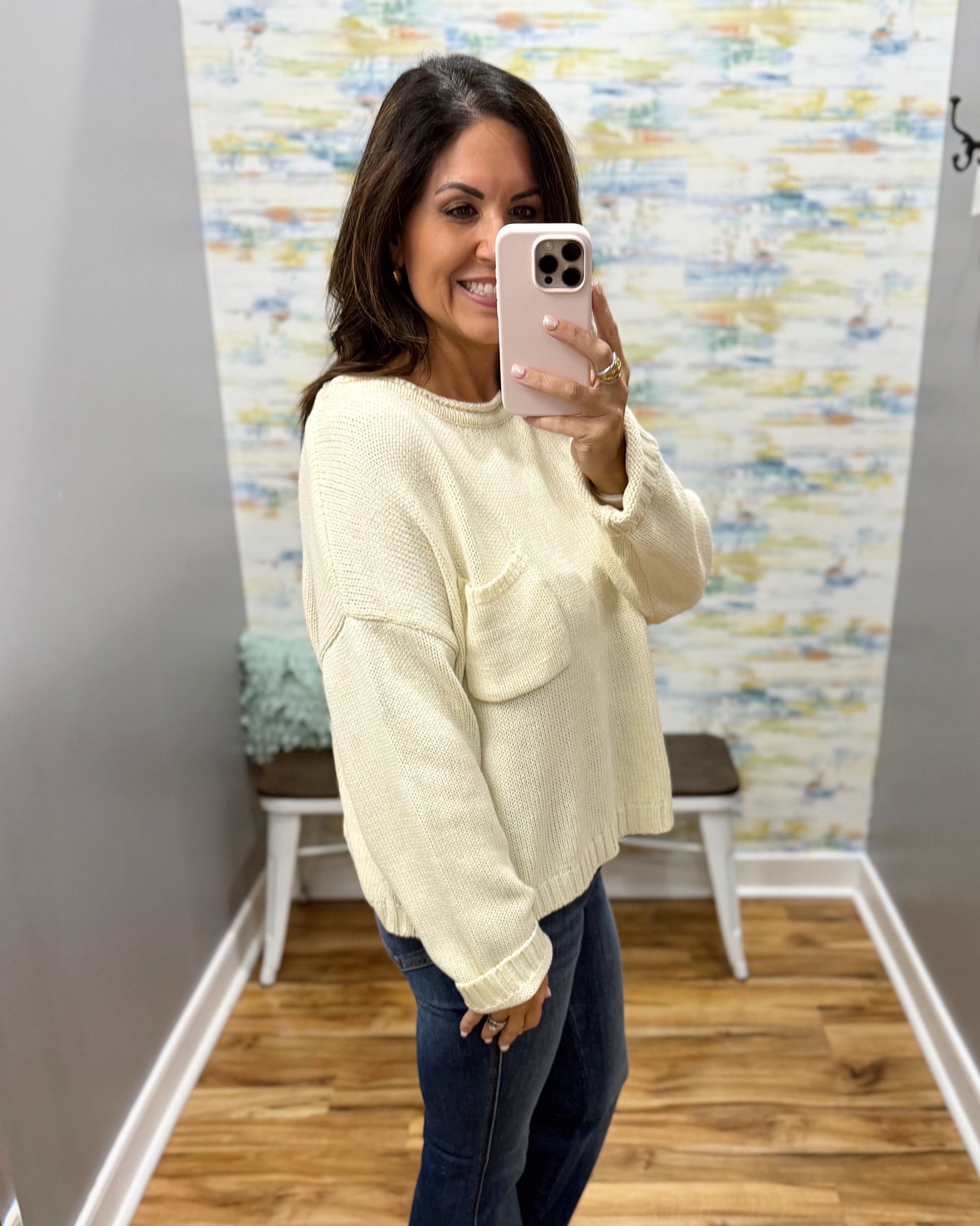 A woman takes a mirror selfie wearing a cream-colored knit sweater with a textured knit pattern and two front patch pockets, paired with dark blue jeans.