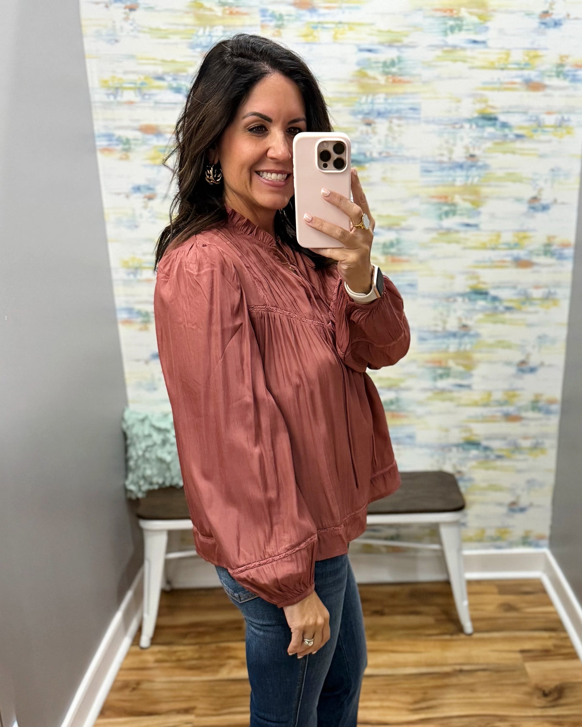 A woman takes a mirror selfie wearing a dusty rose metallic satin jacket and blue jeans in a fitting room with a patterned wallpaper background.