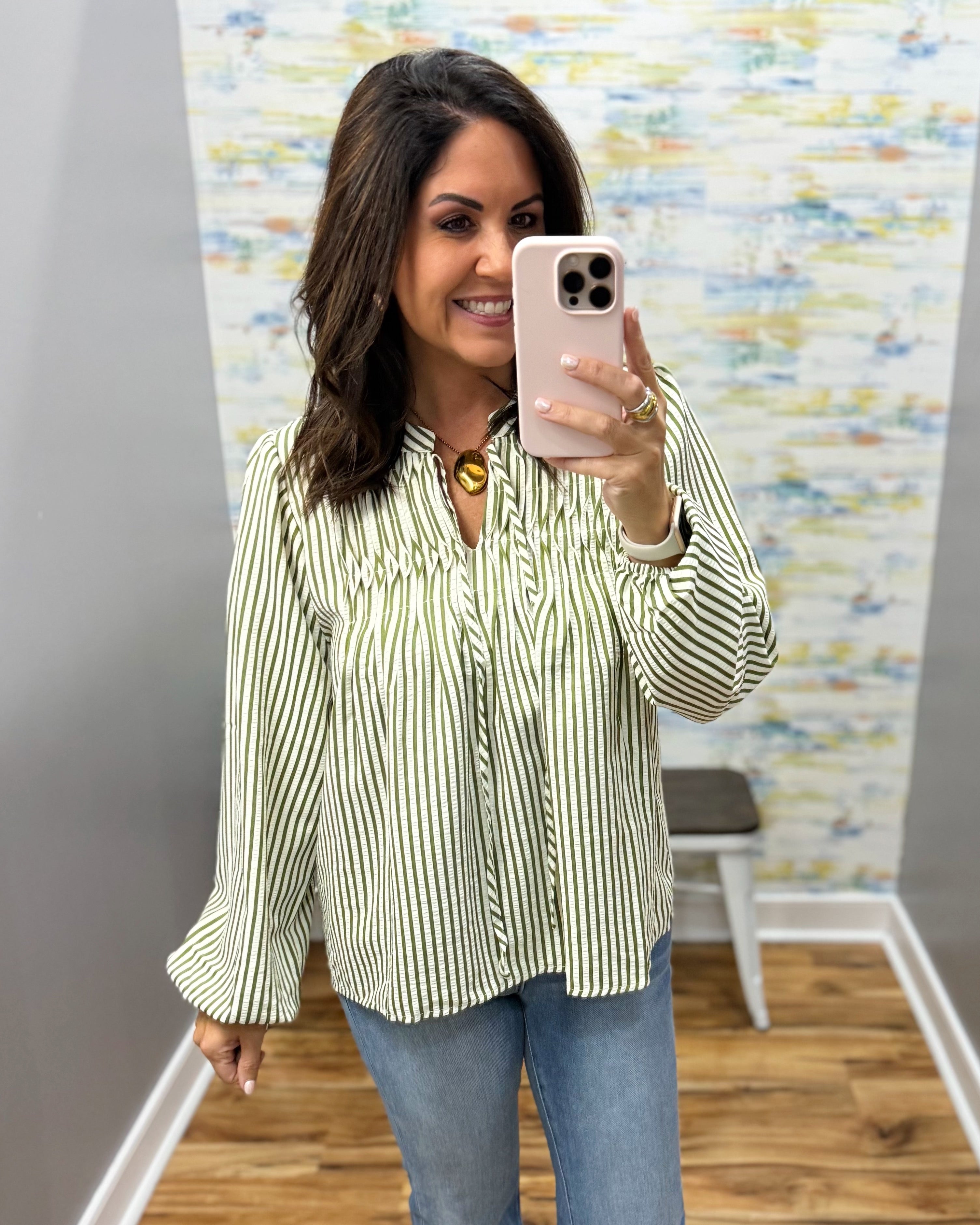 A woman takes a mirror selfie wearing a green and white vertically striped long-sleeve blouse with blue jeans, standing in an indoor room with a patterned wallpaper background.