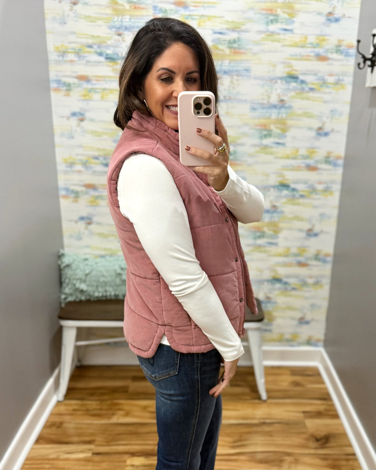 A woman takes a mirror selfie wearing a dusty rose pink quilted vest over a white long-sleeve shirt and blue jeans in a home setting with wood floors and a patterned wallpaper background.