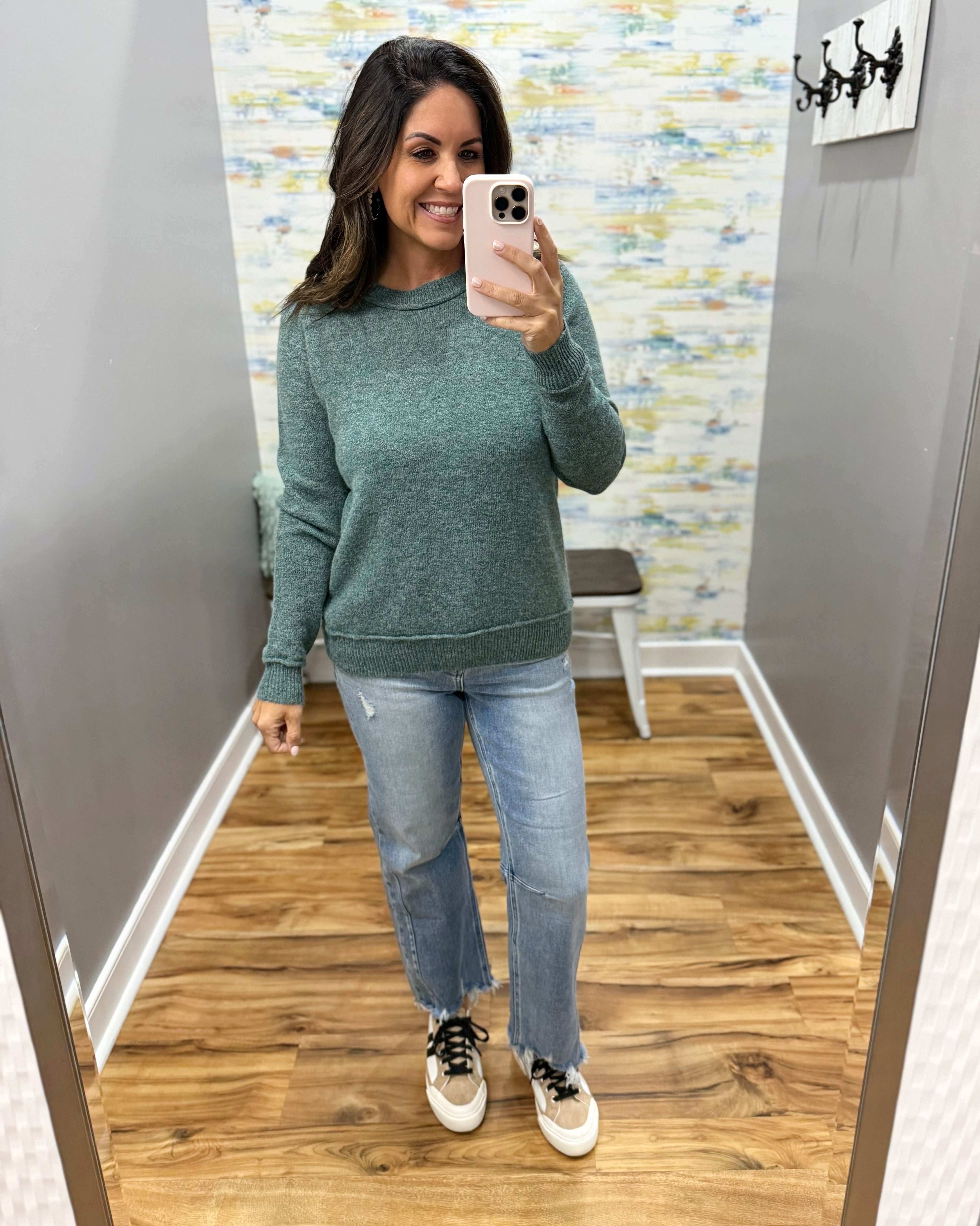 A woman takes a mirror selfie wearing a heathered sage green crewneck sweater, light-wash blue jeans, and white sneakers with black laces in a fitting room.