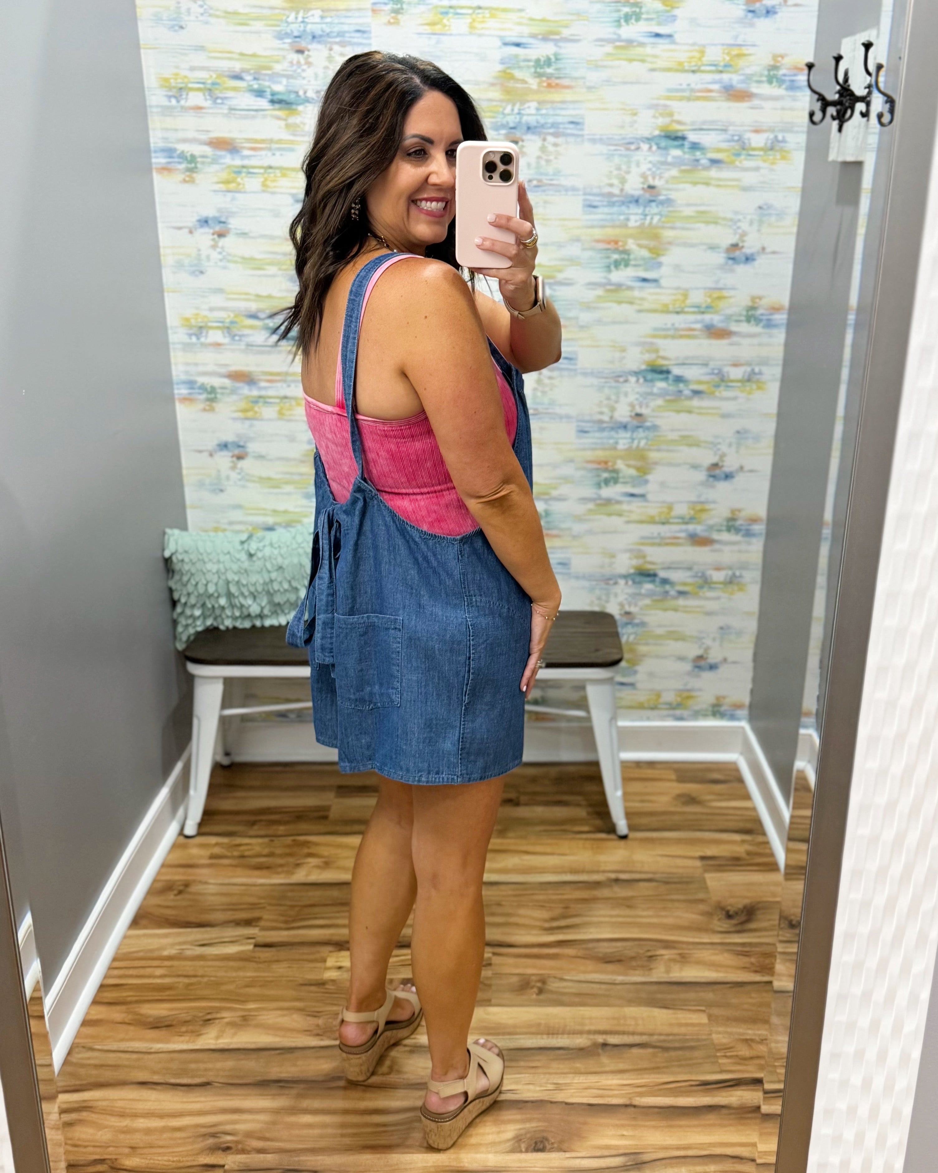 Woman taking a mirror selfie wearing a pink top and blue denim dress in a room with a textured wall.