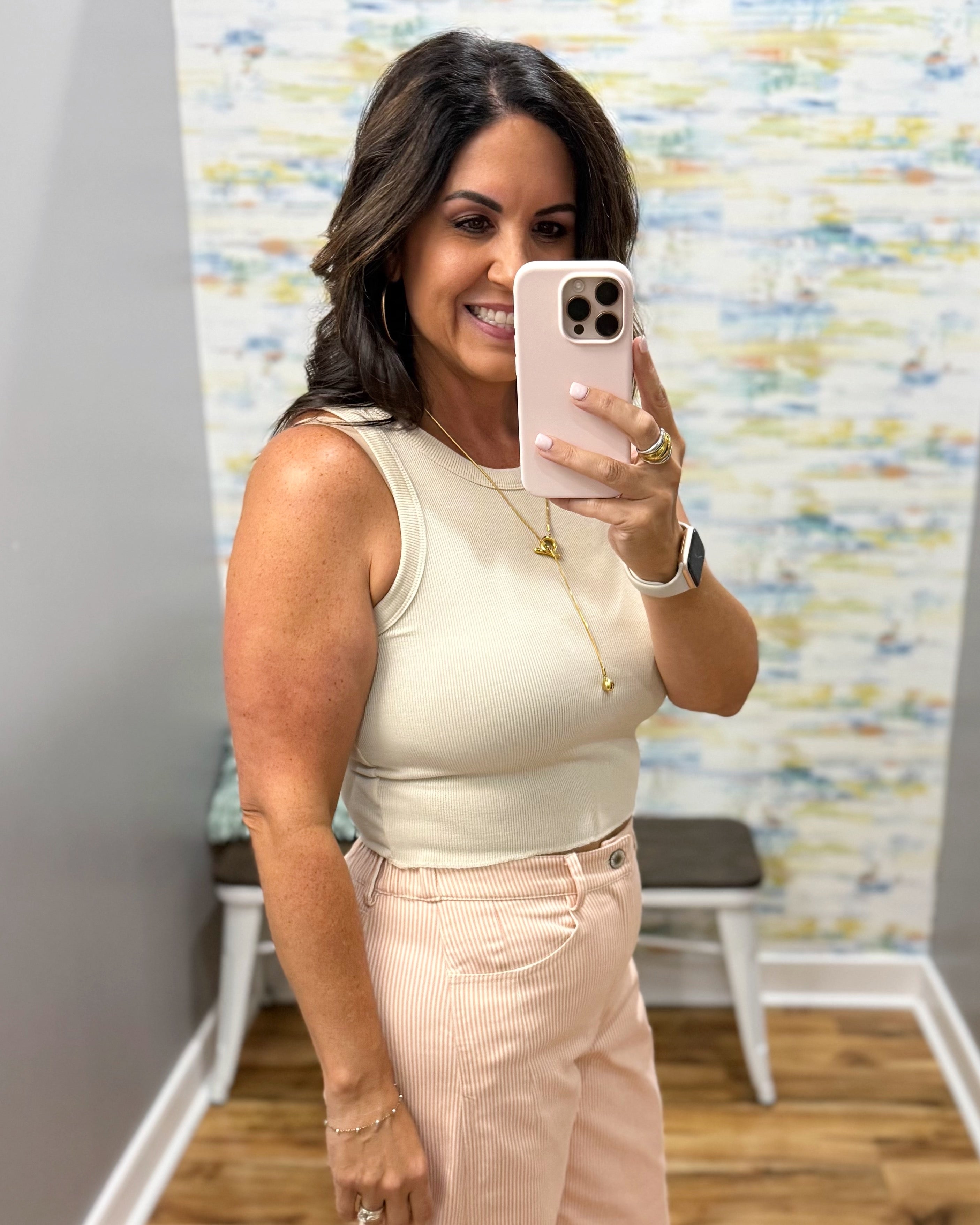 A woman taking a mirror selfie wearing a sleeveless cream top and light pink striped pants, holding a light pink smartphone.