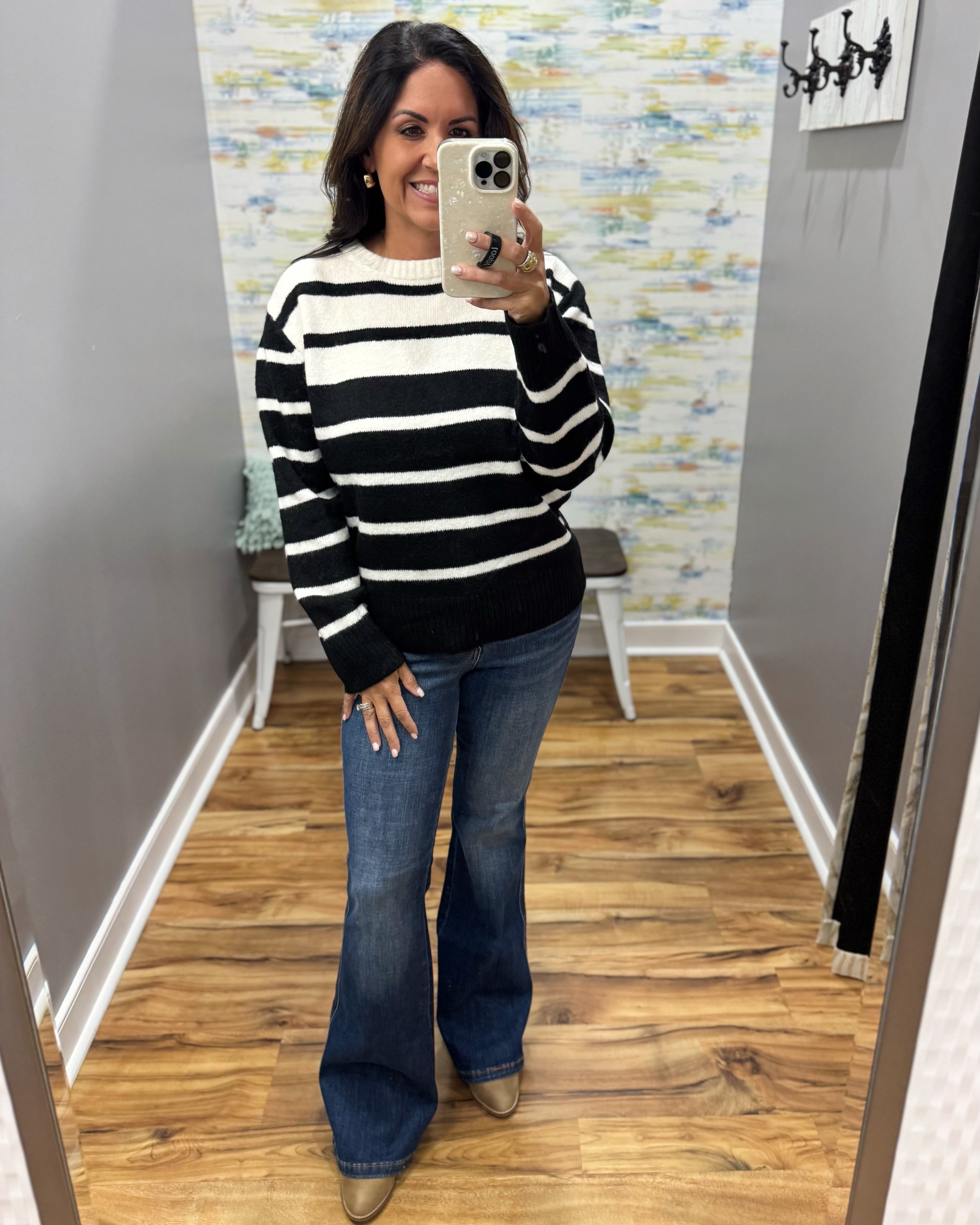 A woman takes a mirror selfie wearing a black and white horizontally striped knit sweater, blue flare jeans, and tan pointed-toe flats in a home setting with wood floors and gray walls.