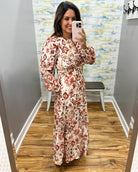 A woman takes a mirror selfie in a fitting room wearing a long-sleeve maxi dress with a rust red and beige geometric pattern on a cream background.