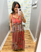 A woman takes a mirror selfie in a home hallway wearing a sleeveless, tiered maxi dress with a red-orange base and a multicolor patchwork print featuring floral and paisley patterns.