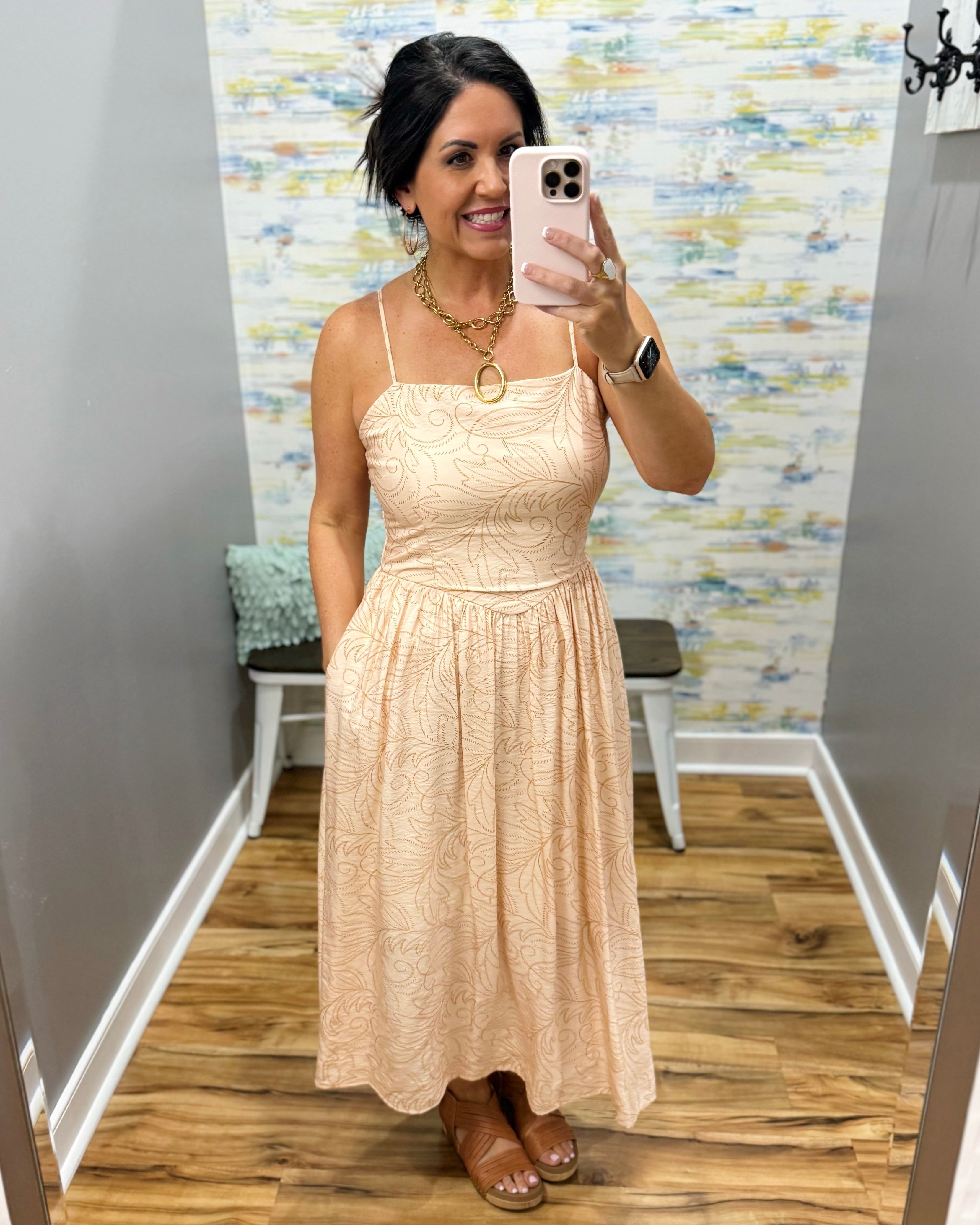 Woman taking a mirror selfie wearing a beige lace dress in a room with a patterned wall.