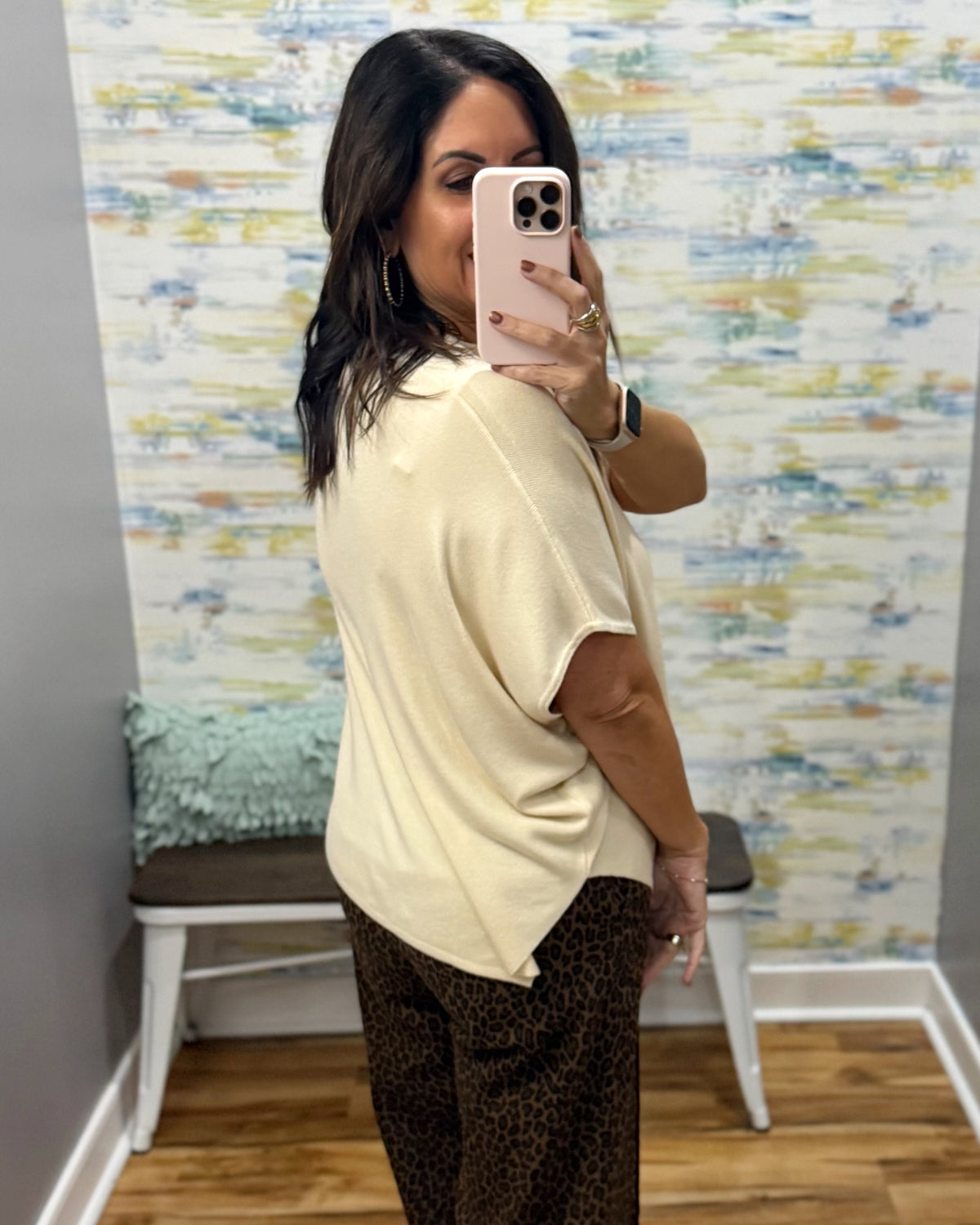 A woman takes a mirror selfie in a fitting room wearing a cream short-sleeve knit top and brown leopard-print pants. The background features a colorful abstract wallpaper and a small bench.