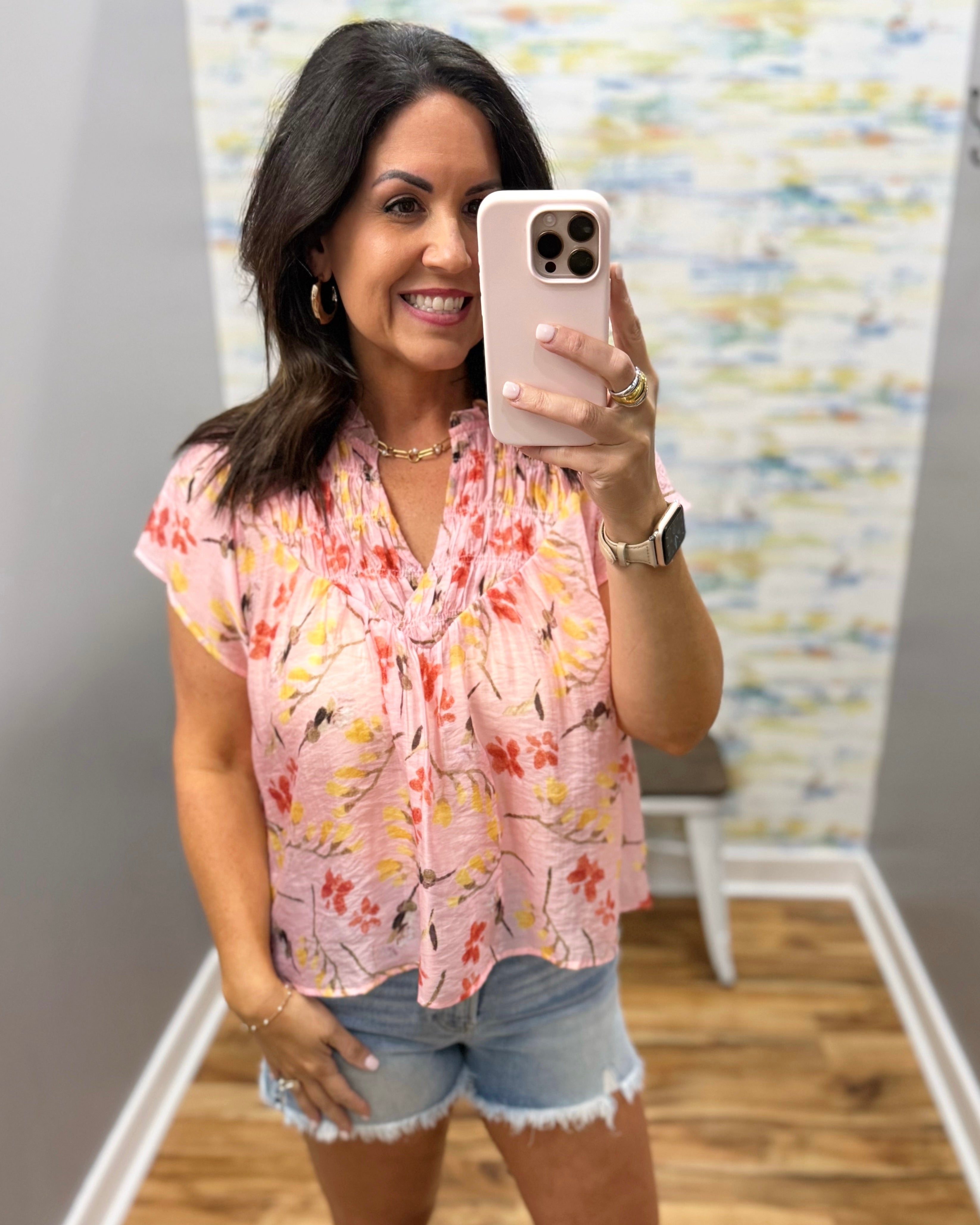 Woman taking a mirror selfie wearing a floral blouse and denim shorts in a room with a colorful wall.