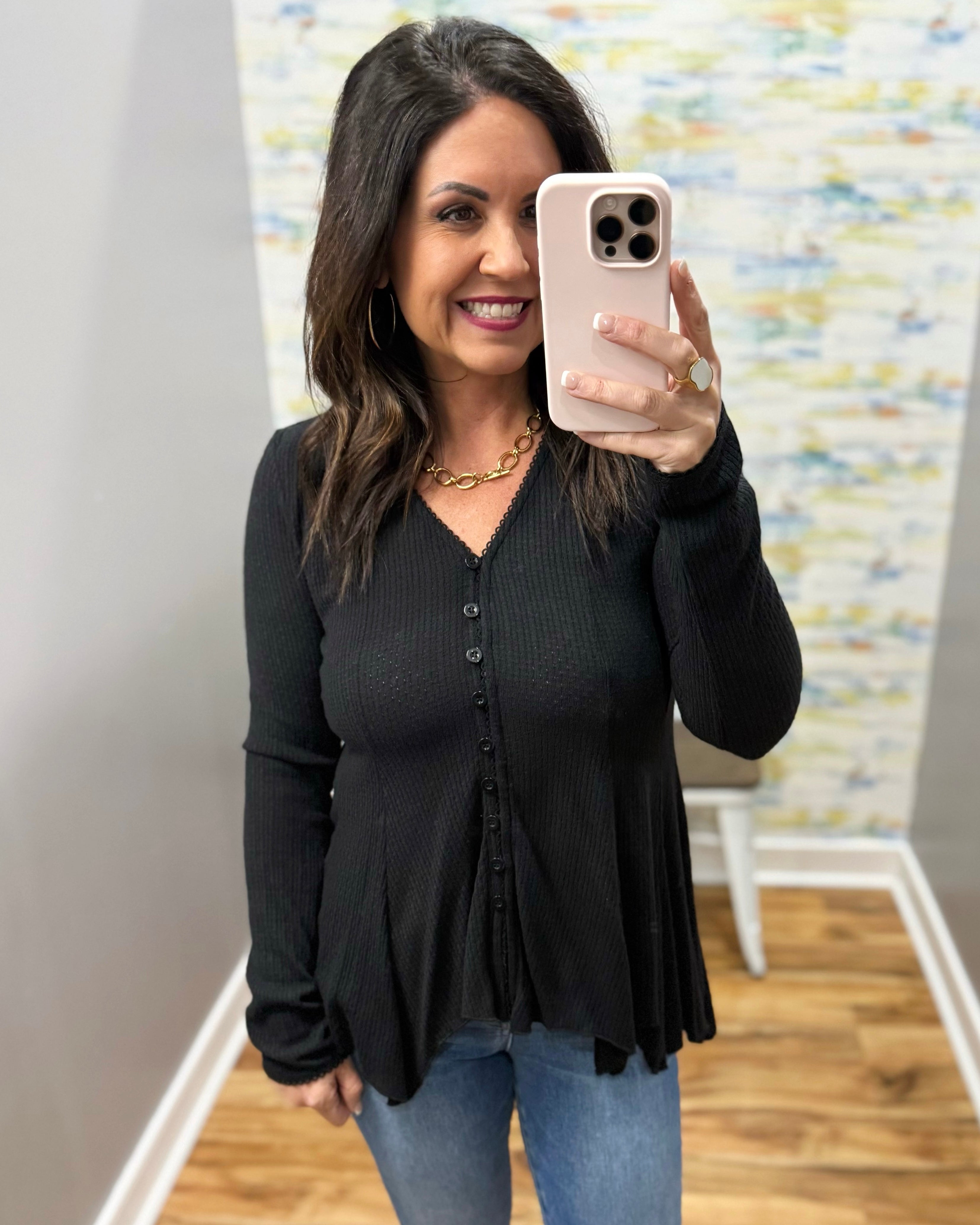 Woman taking a mirror selfie wearing a black long-sleeve top and blue jeans, holding a phone.