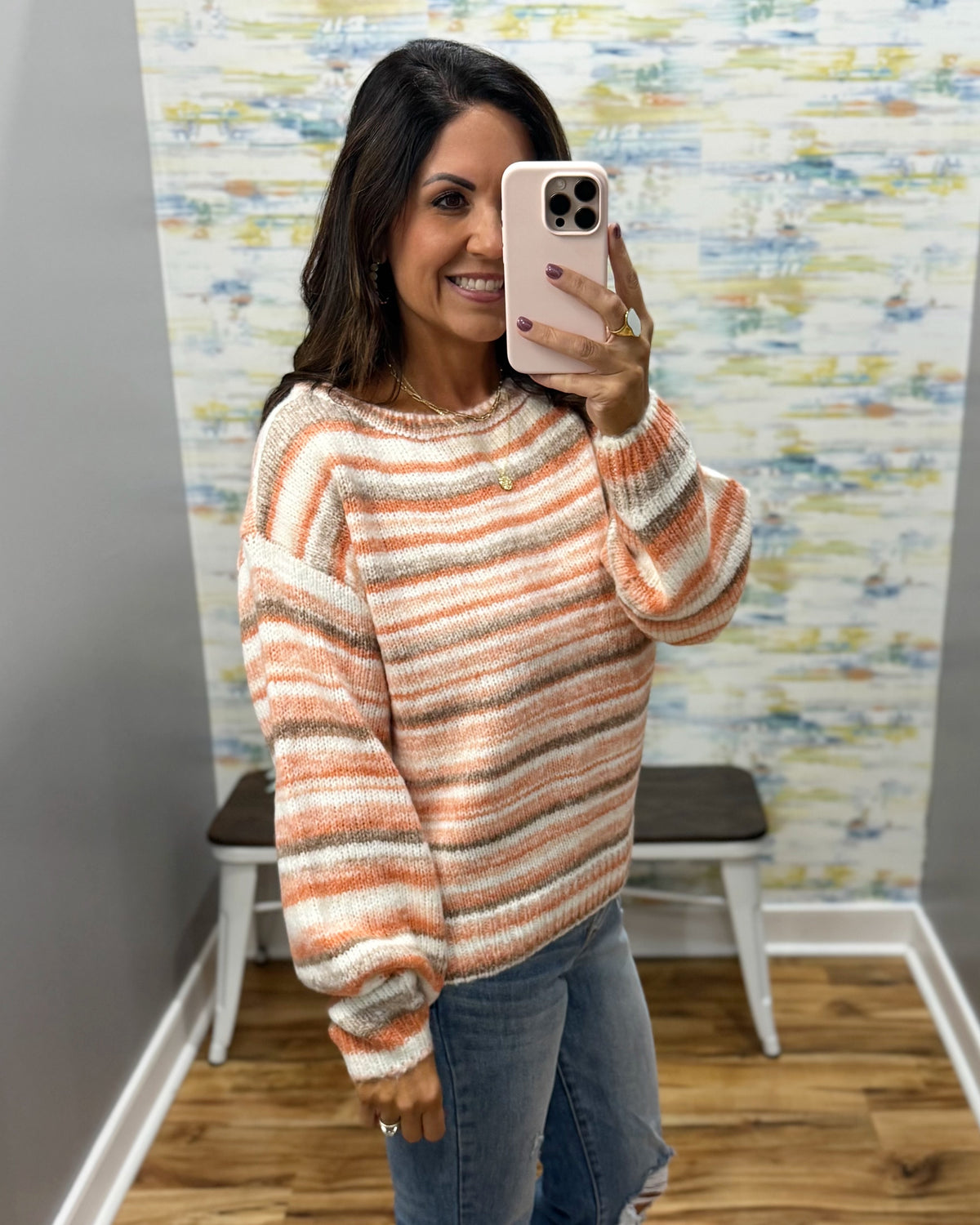 A woman takes a mirror selfie wearing a fuzzy, textured knit sweater with horizontal stripes in orange, beige, and white. She is also wearing blue jeans, standing in front of a patterned wallpaper background.