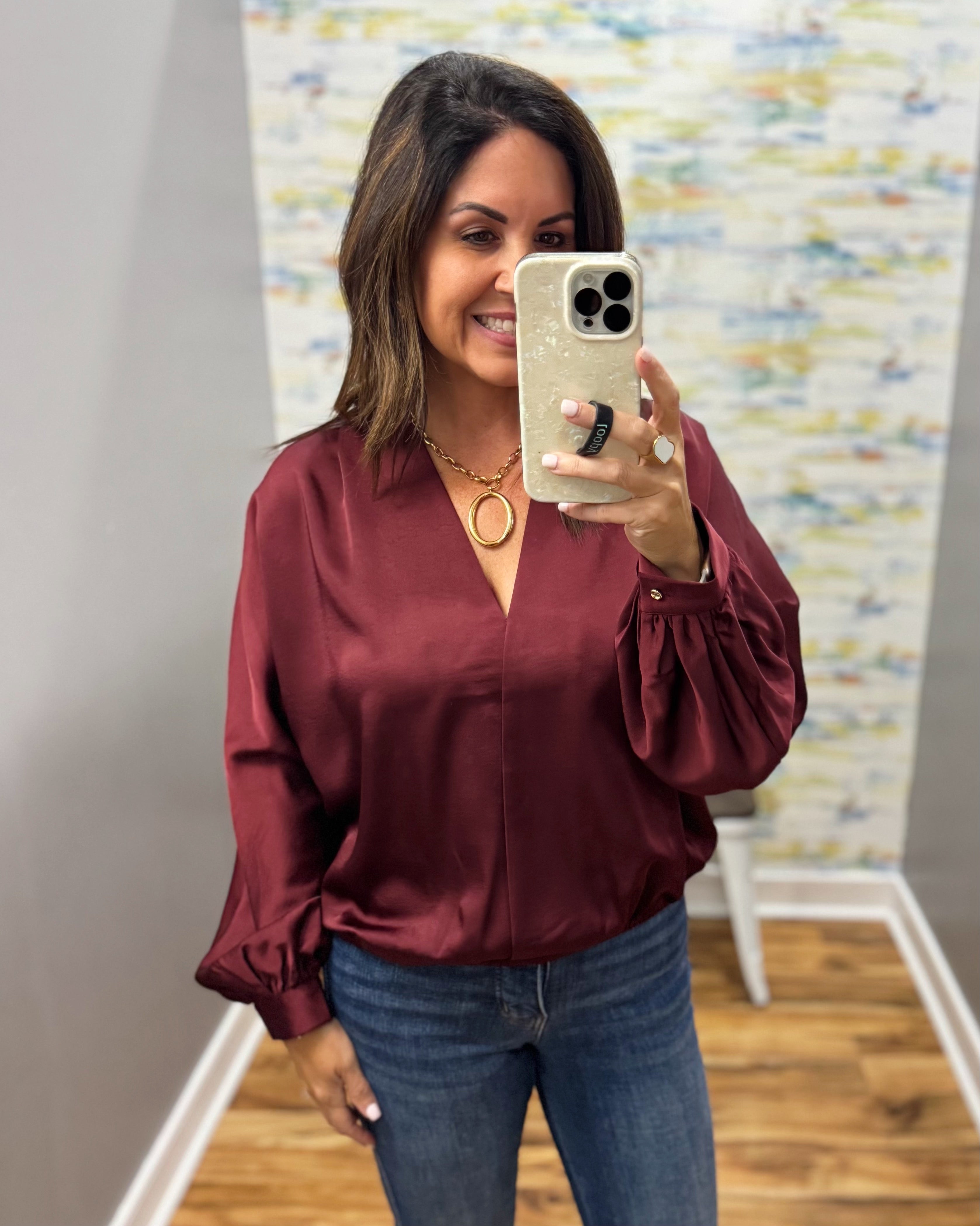 A woman takes a mirror selfie wearing a long-sleeve satin blouse in deep burgundy with blue jeans. The blouse features a V-neckline and gathered sleeves.