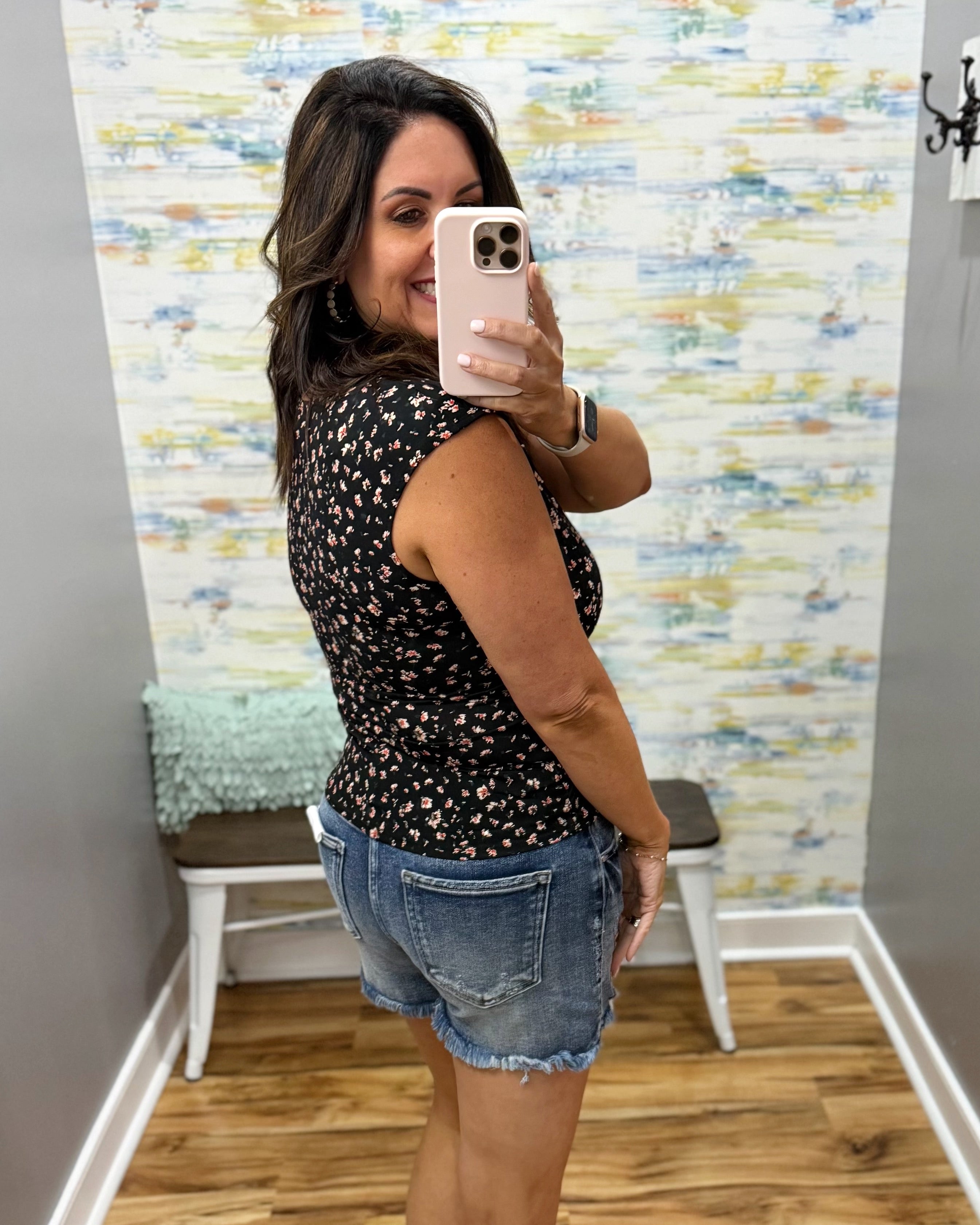 Woman taking a mirror selfie wearing a black floral top and denim shorts in a room with colorful wall paper.