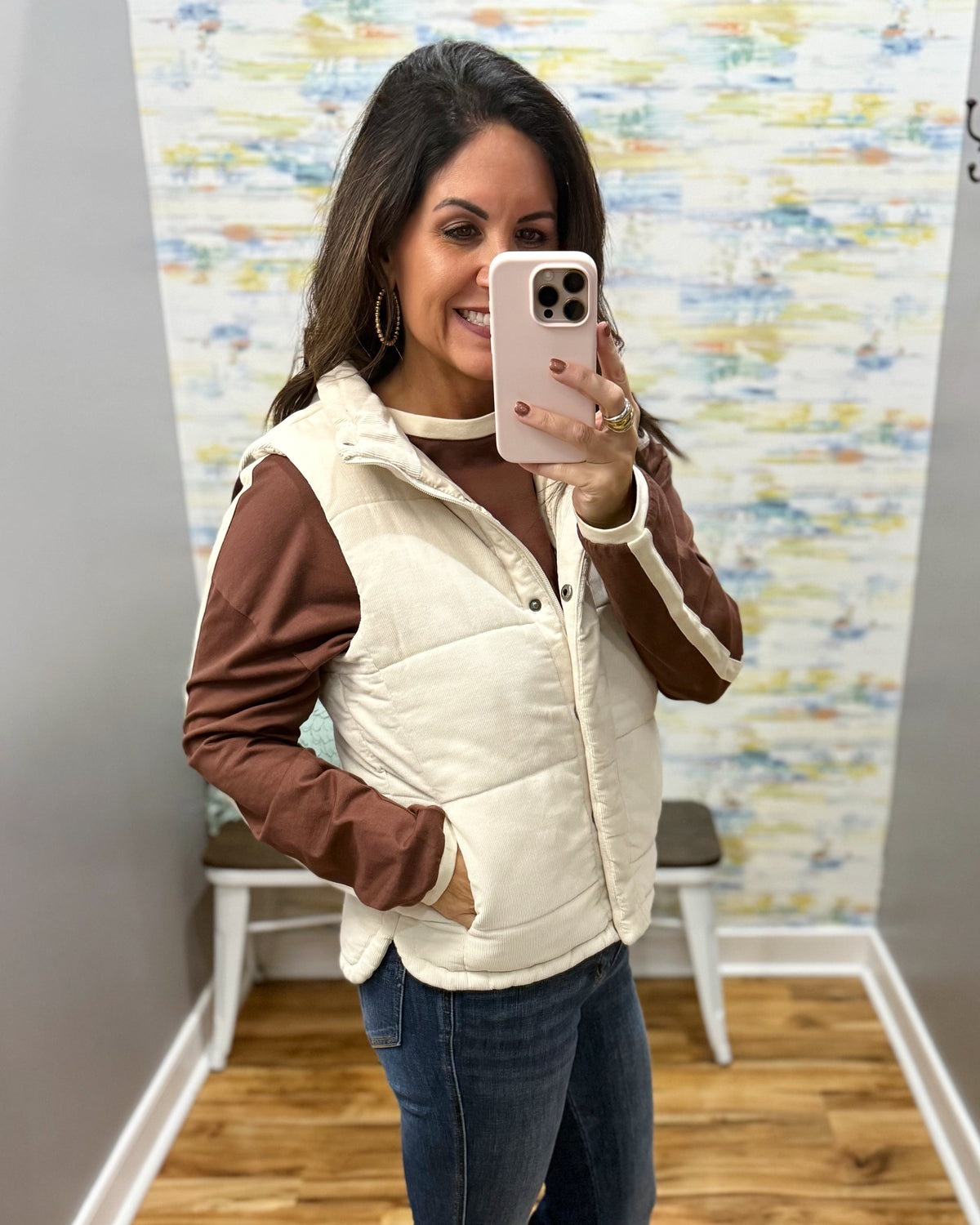 A woman takes a mirror selfie wearing a cream-colored puffer vest over a brown long-sleeve shirt and blue jeans. The setting appears to be a boutique fitting room with a patterned wall and wood floor.