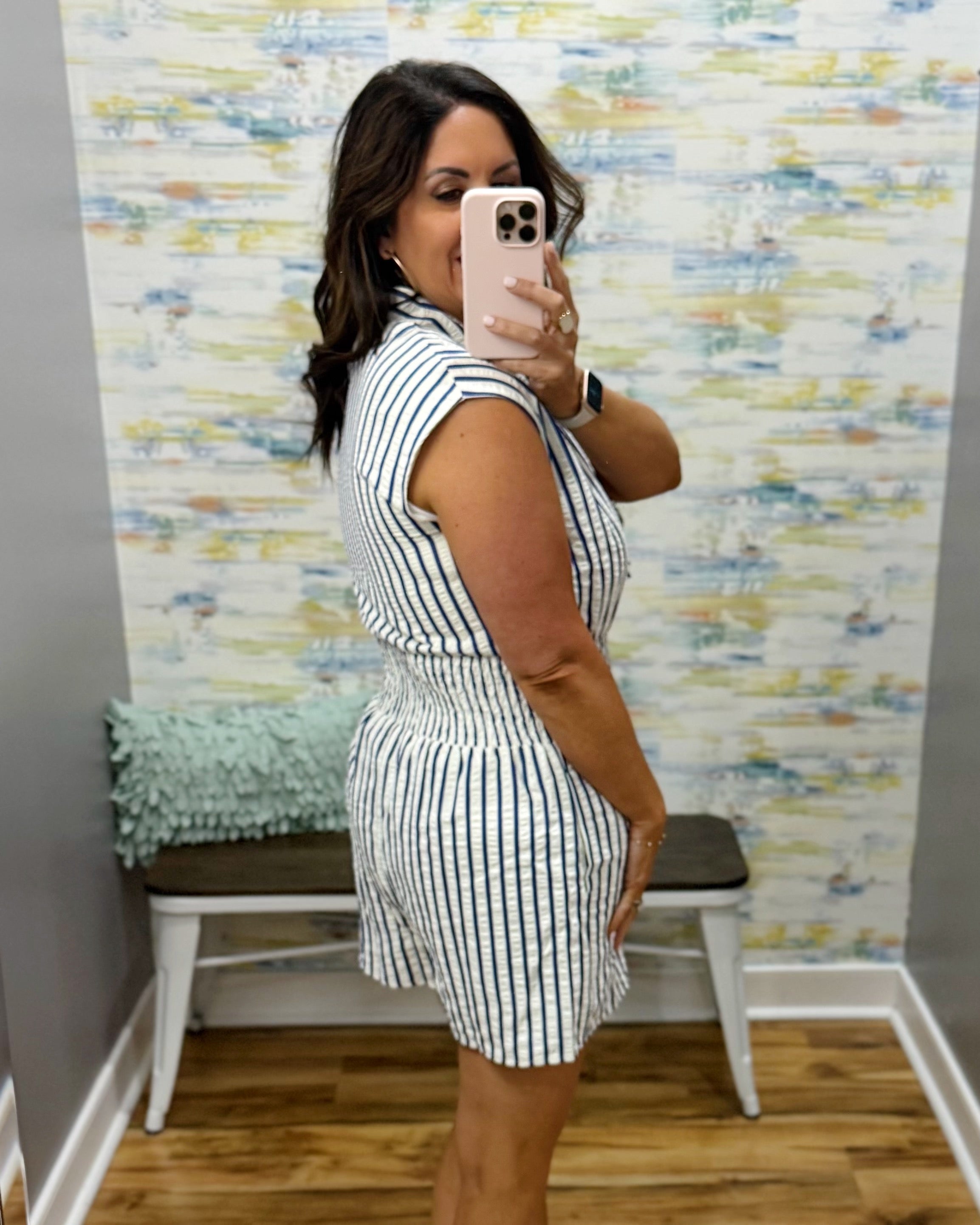 Woman taking a mirror selfie wearing a striped romper in a room with a colorful wall.