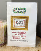 A packaged cross stitch kit in a clear plastic sleeve. The front shows a white card with a framed sample cross stitch design reading 'HOT GIRLS HAVE GRANDMA HOBBIES' and the brand name 'subversive cross stitch' at the top.