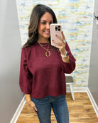 A woman takes a mirror selfie wearing a heathered burgundy knit sweater, blue jeans, and gold jewelry in a home setting with a colorful abstract wallpaper.