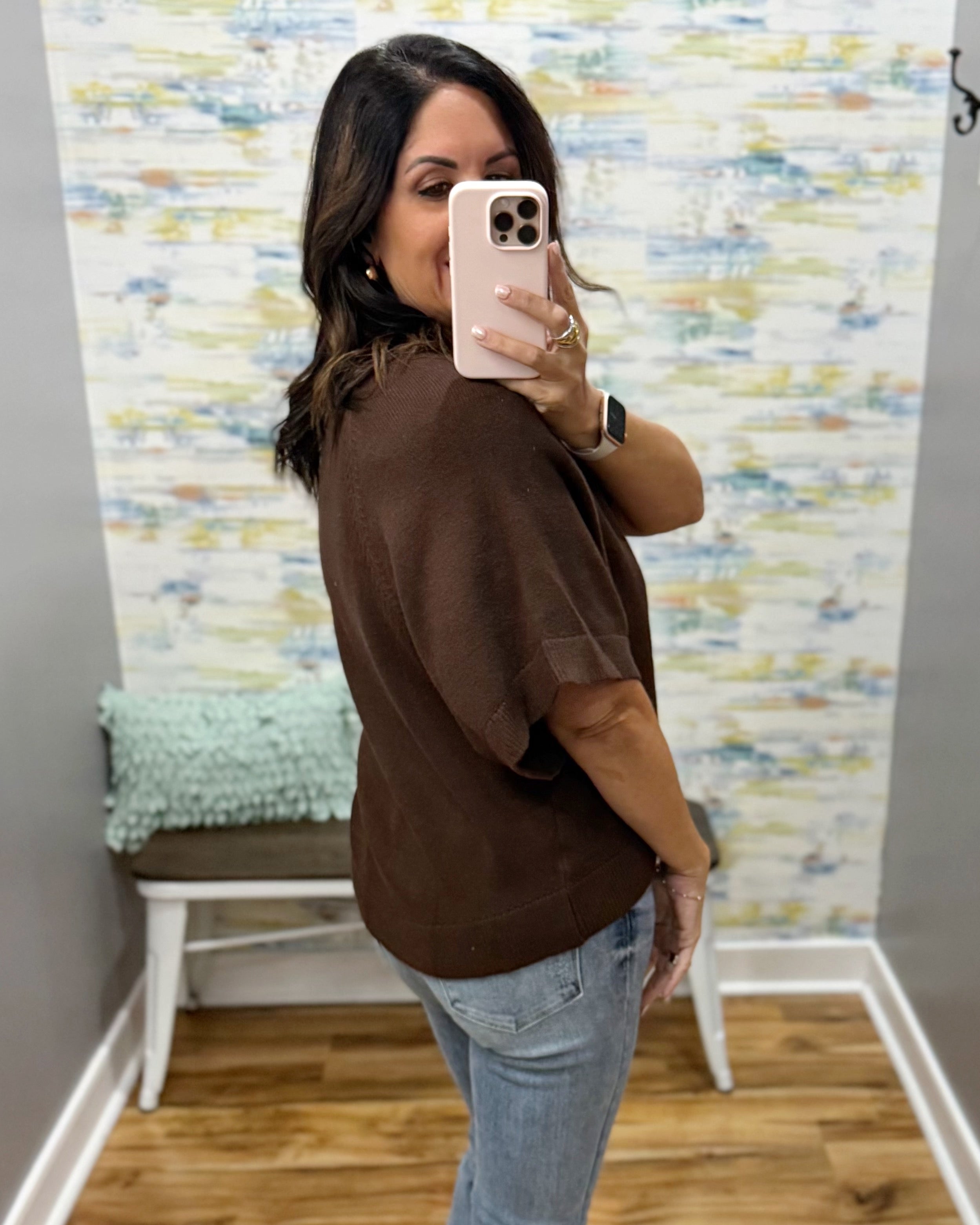 A woman takes a mirror selfie wearing a brown short-sleeve top and light blue jeans in a fitting room with a colorful abstract wallpapered wall.
